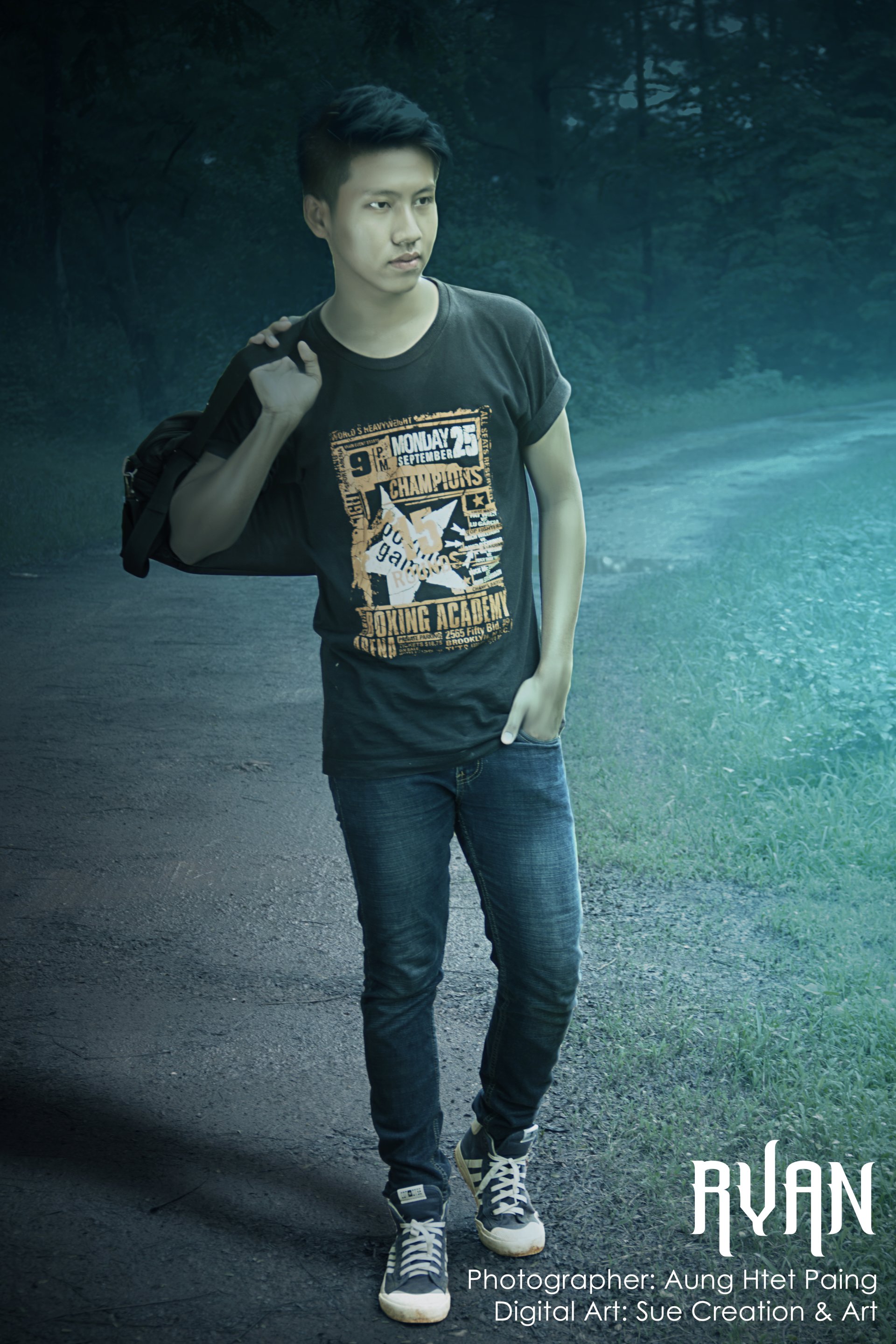 An Asian man stands on a path surrounded by greenery, carrying a bag over his shoulder and wearing a graphic T-shirt and jeans.