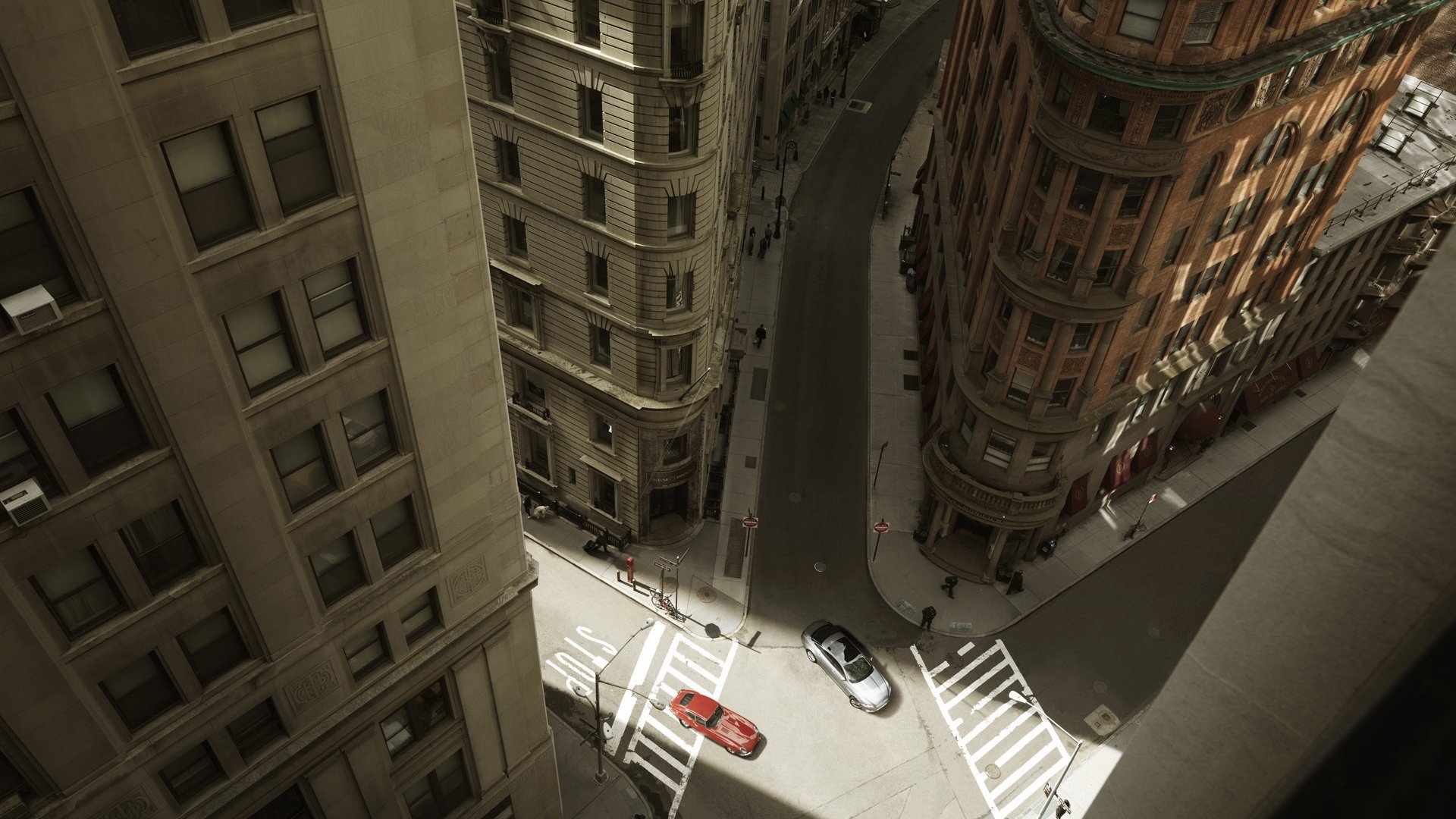 A bird's-eye view of a man-made urban road flanked by tall buildings with a few cars navigating the intersection below.