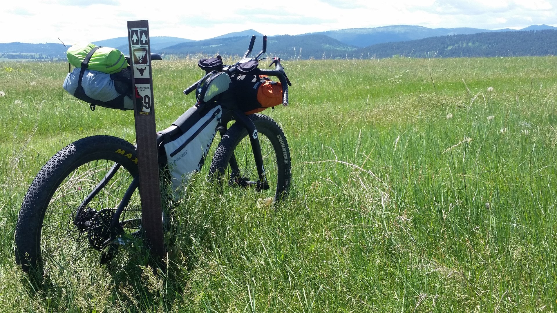 A bicycle stands in a grassy field, with a travel sign nearby and mountains in the background, showcasing a serene outdoor adventure setting.