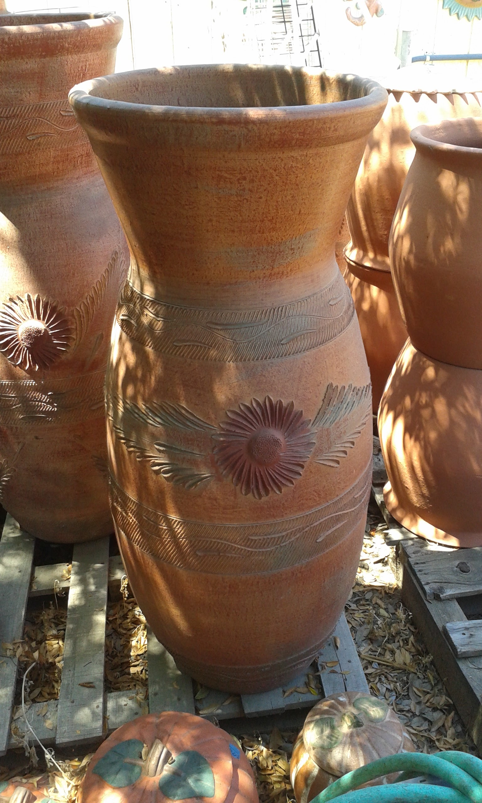 Tall man-made terracotta pottery vase with carved flowers and wave motifs, standing among other clay pots on wooden pallets outdoors.