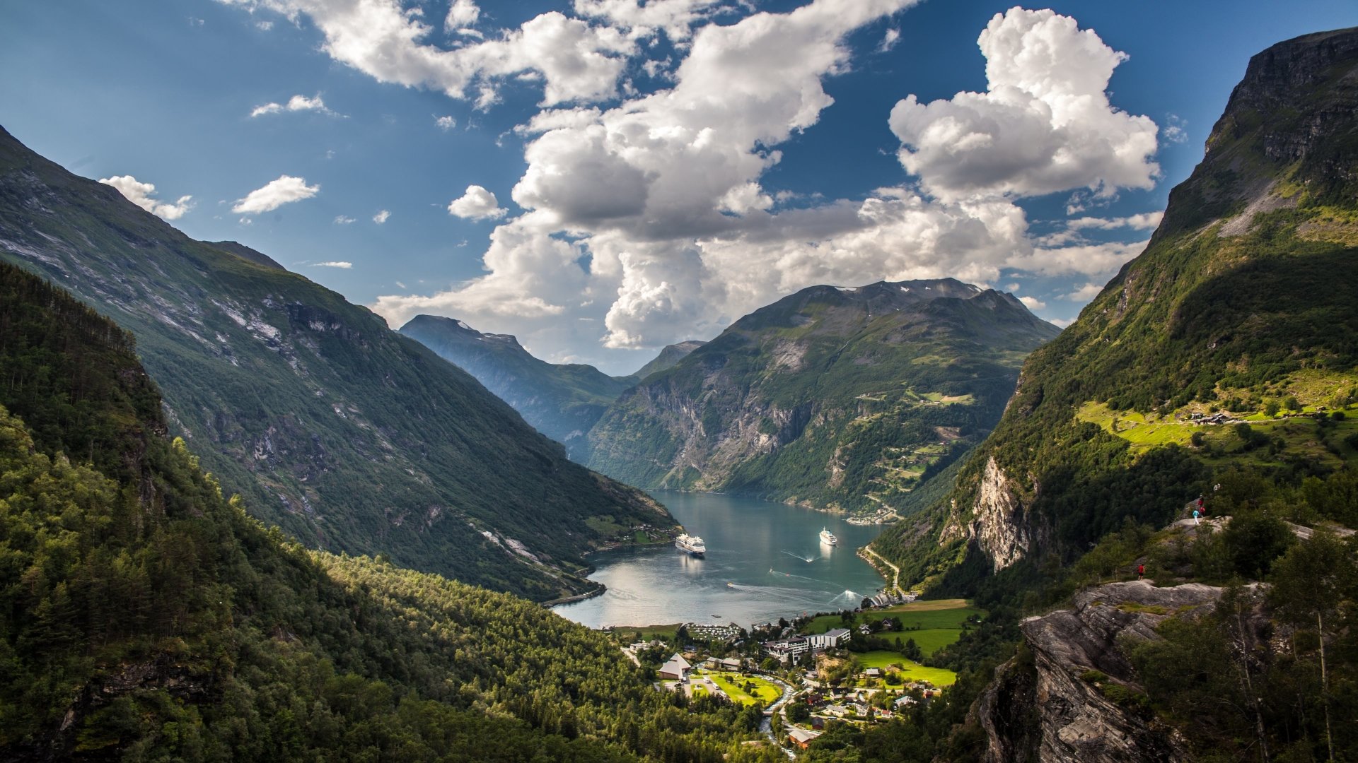 Landscape photography of a sunlit fjord valley with steep green mountains, a small village on the shore and a cruise ship under a dramatic cloud-filled sky.