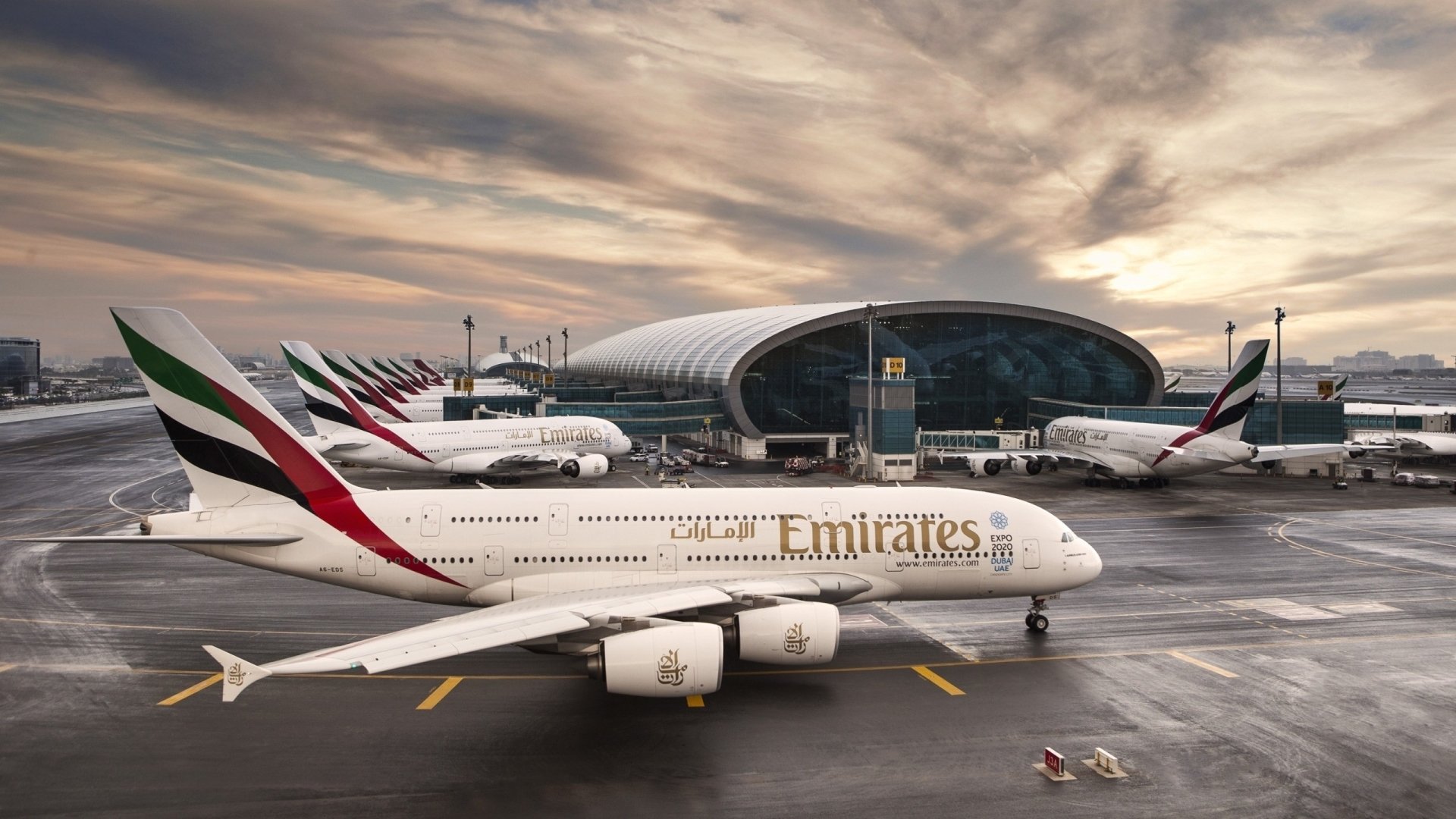 An Emirates Airbus A380 is parked on the tarmac at an airport, with multiple aircraft and a modern terminal building in the background under a dramatic sky.