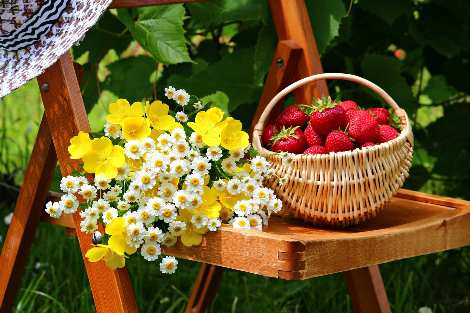 A wooden shelf displays a basket of ripe strawberries beside a bouquet of yellow and white flowers, creating a vibrant still life composition in a lush outdoor setting.