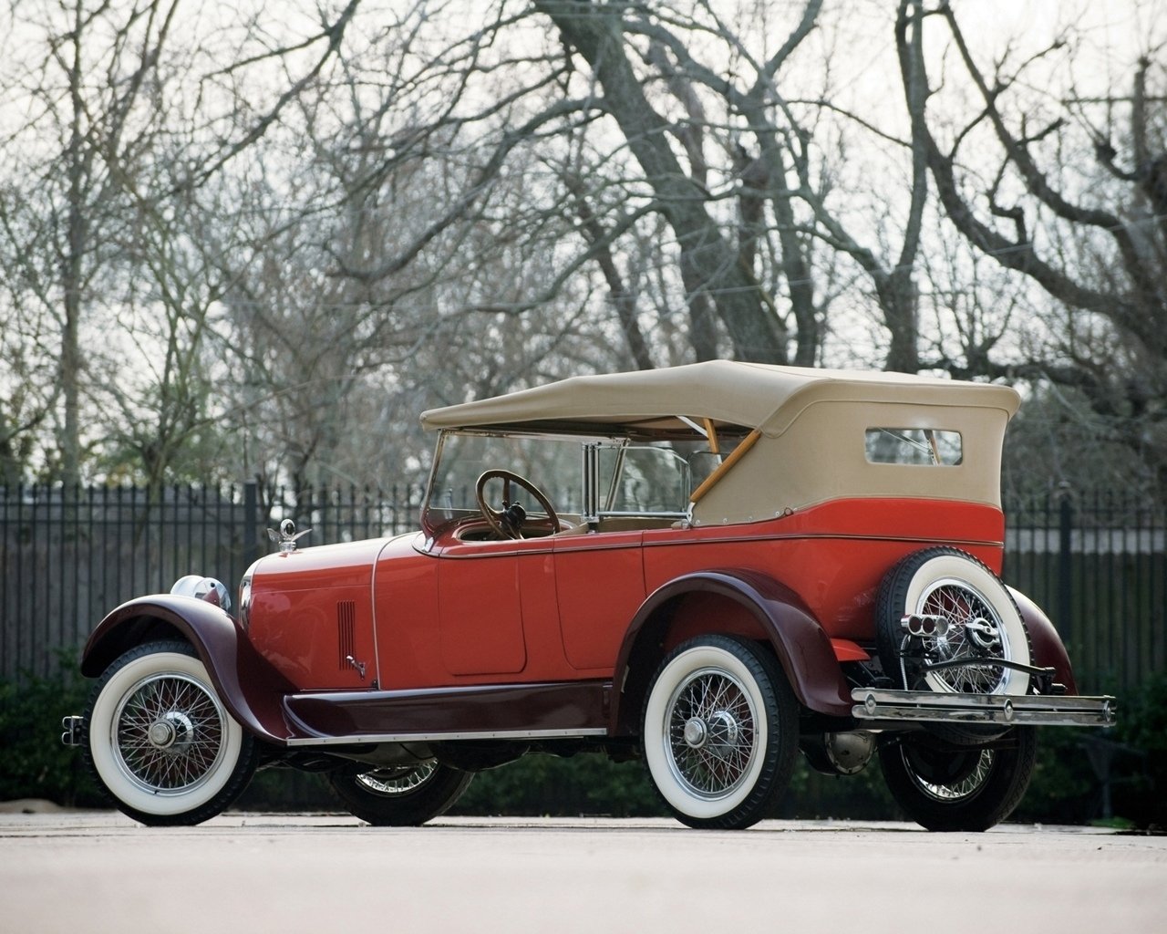 Red and tan Duesenberg Model A Phaeton convertible parked roadside, classic 1920s styling with whitewall tires, wire wheels and rear-mounted spare.