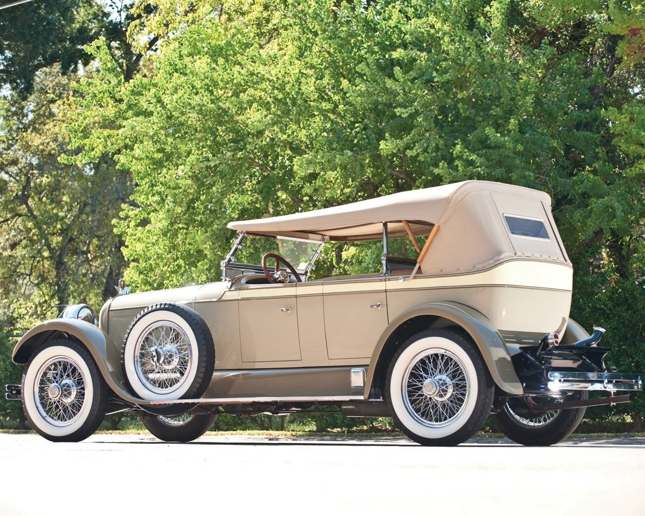 Beige Duesenberg Model A Phaeton convertible parked roadside, side view with dual side-mounted spare wheels, whitewall tires and tan folding top.
