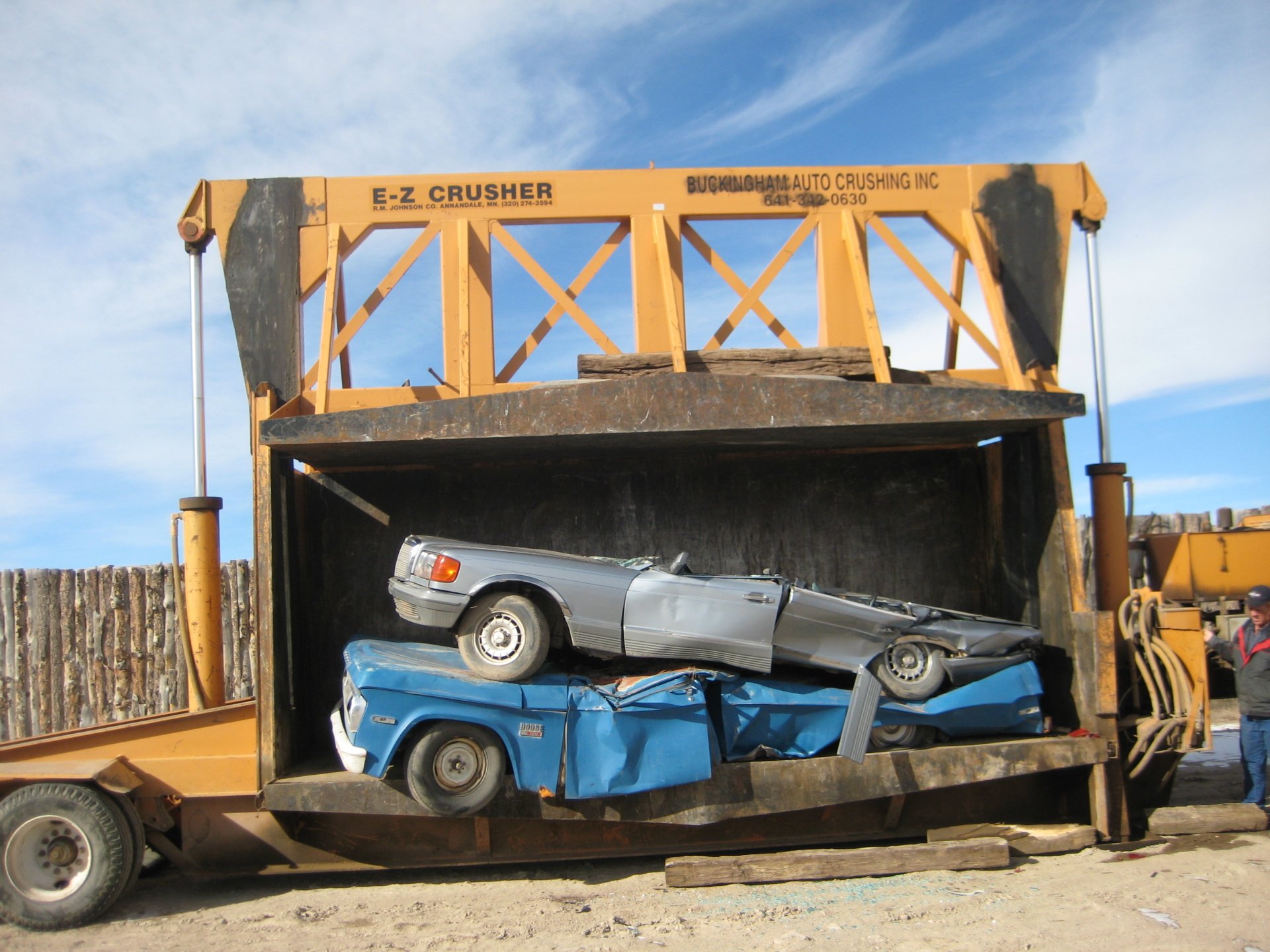 A wrecked vehicle is crushed inside a large machine, with a silver car atop a blue one, amidst a clear blue sky and industrial surroundings.