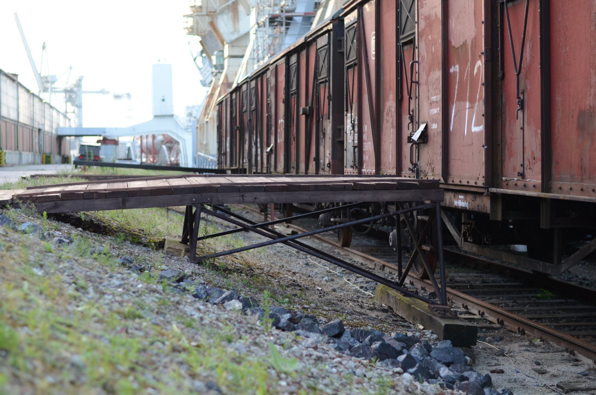Rusty freight train cars and rail vehicles parked on a siding beside a raised loading platform, with gravel ballast and industrial buildings in the background.