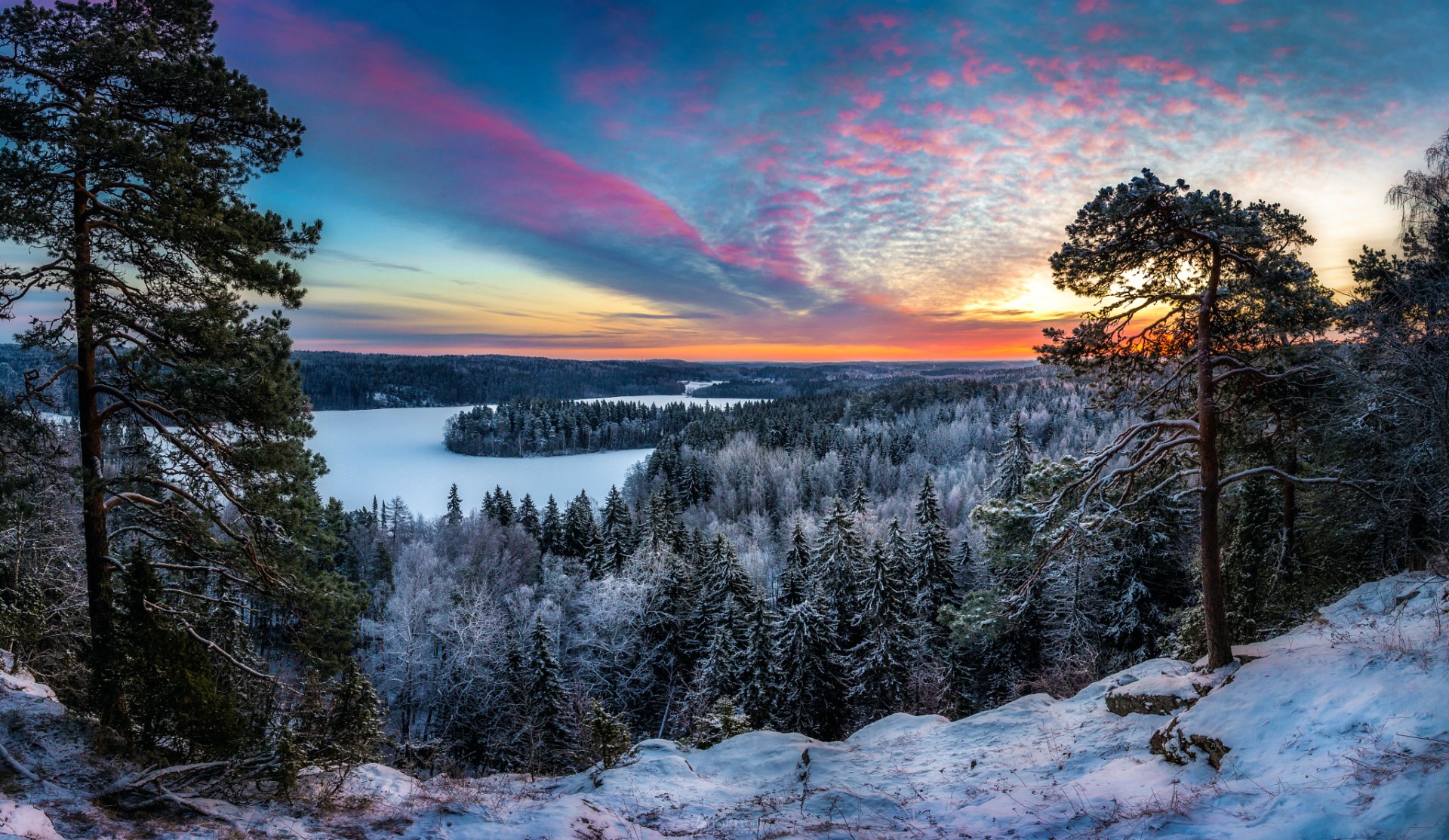A stunning winter sunset over a snowy forest, with vibrant colors reflected in a serene lake, surrounded by tall trees and a blanket of snow covering the landscape.