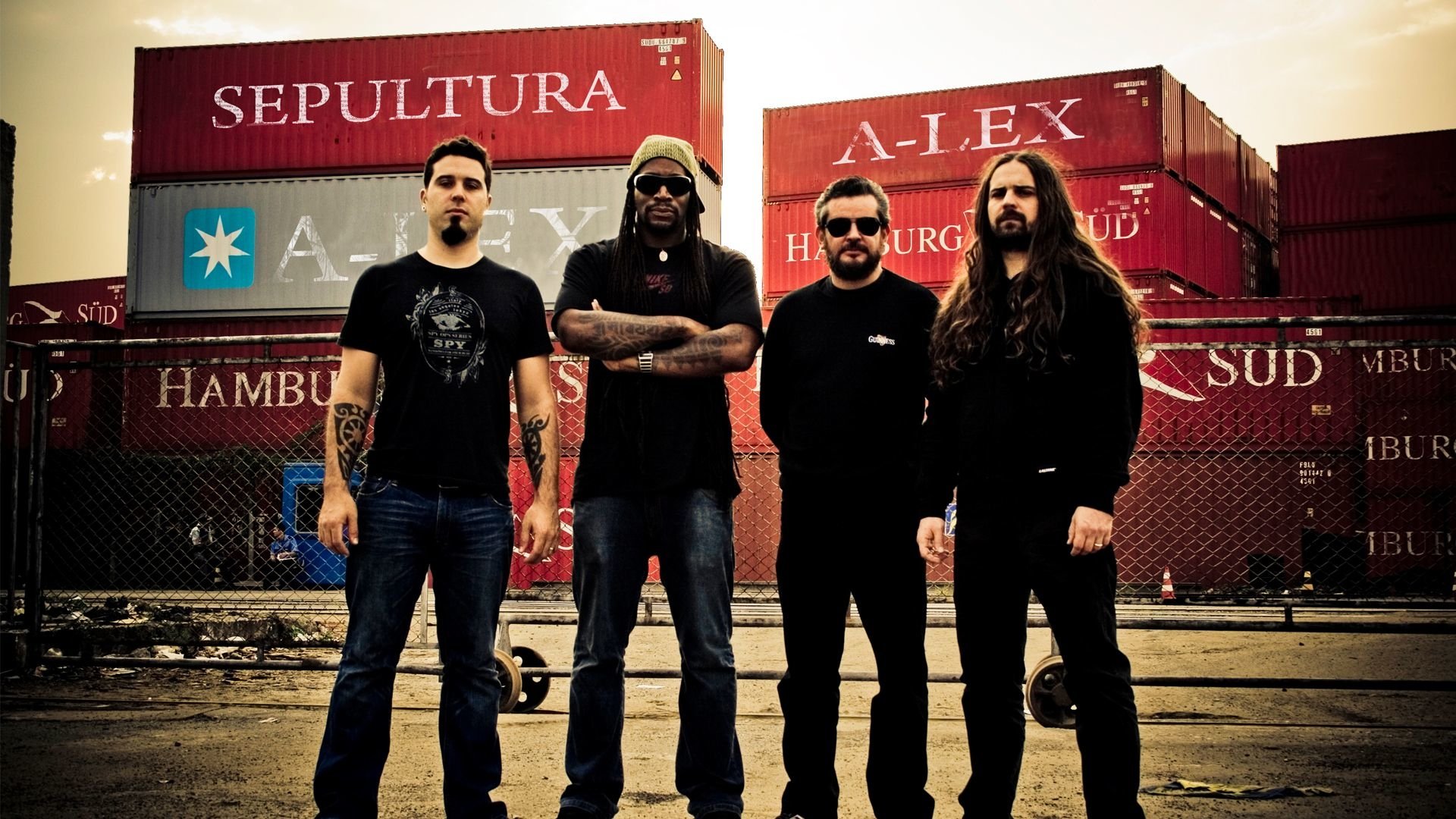Four members of the band Sepultura pose confidently in front of colorful shipping containers, showcasing their music roots and strong presence.
