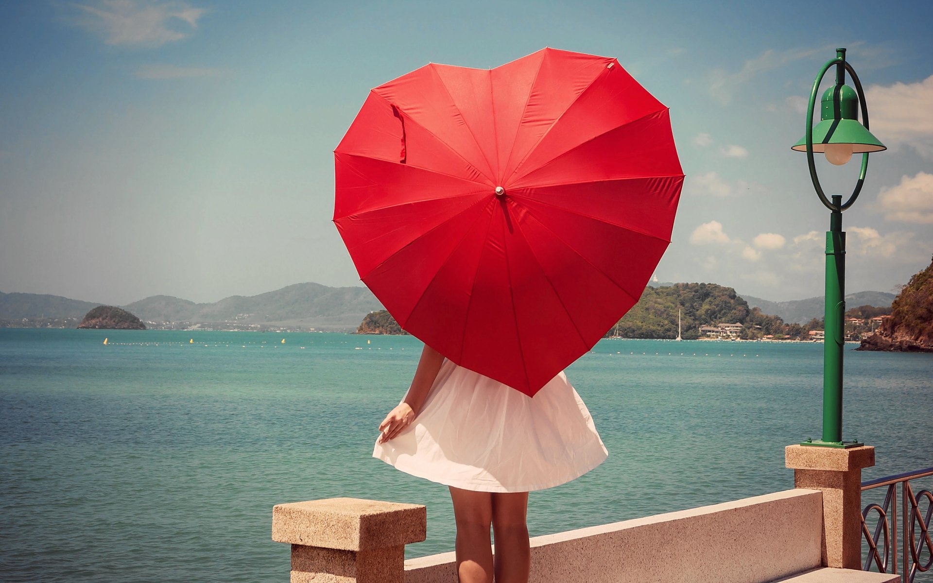 A woman stands by the waterfront holding a red heart-shaped umbrella, with her back facing the camera and a scenic ocean view in the background.