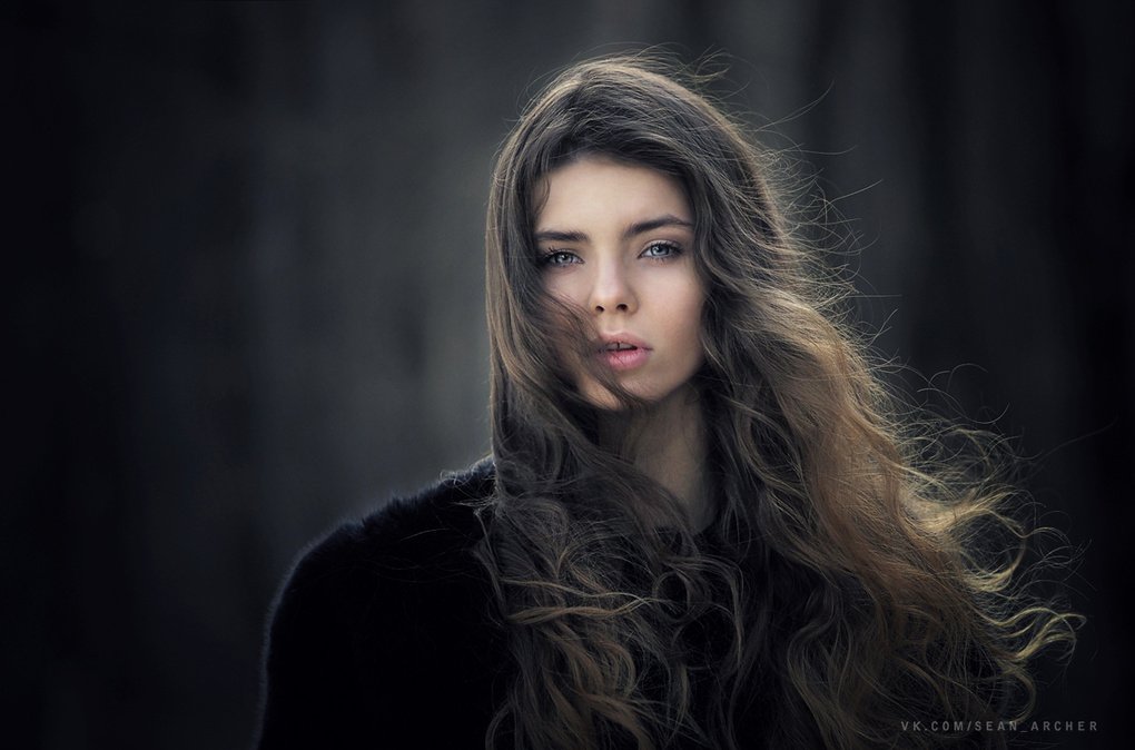 A woman with long, flowing hair gazes confidently into the camera, surrounded by a softly blurred, dark background, showcasing her striking features as a model.