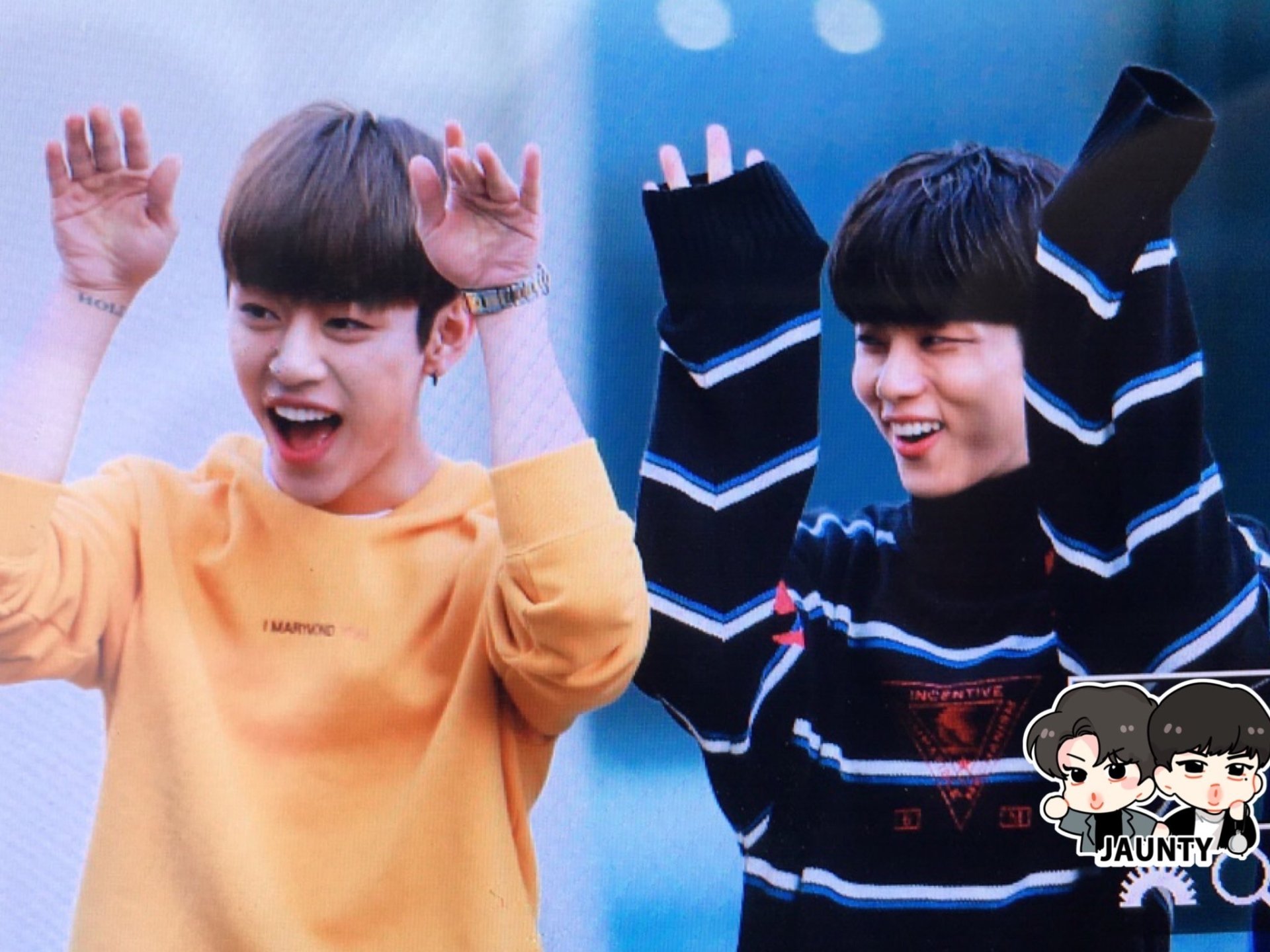 Two young men are joyfully posing with raised hands, wearing colorful sweaters, in a lively outdoor setting. The image reflects the energetic spirit of B.A.P. and their music.