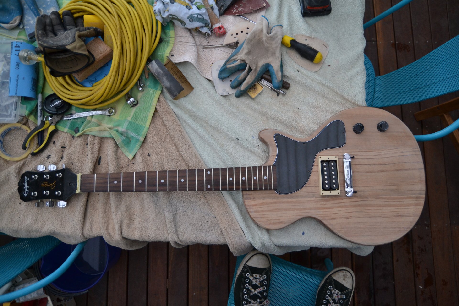 An unfinished electric guitar lies on a cloth-covered surface surrounded by work gloves, tools, and a coiled yellow cable, blending music craftsmanship with a workshop setting.