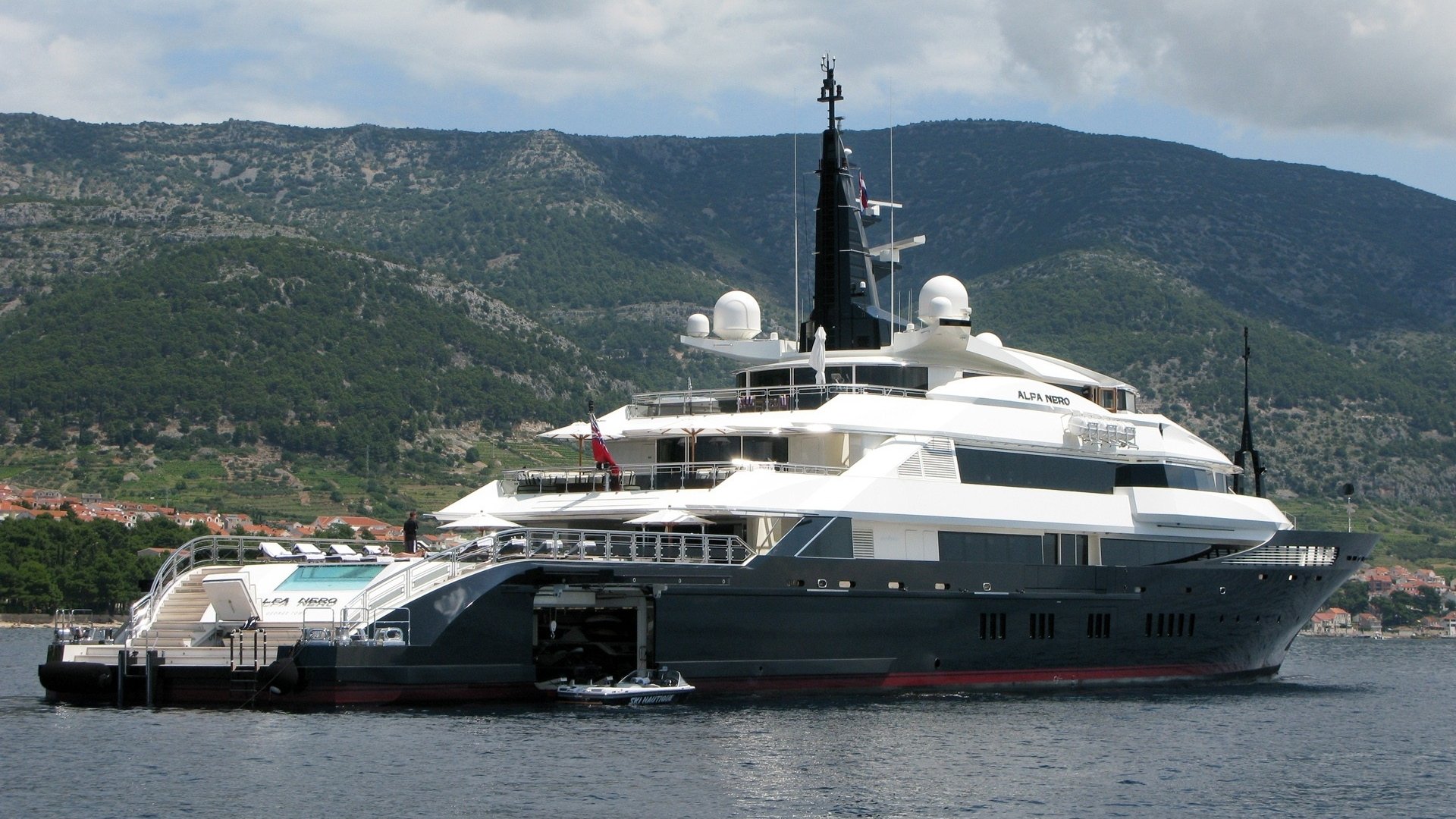 A luxurious yacht with a sleek design is anchored on calm waters, set against a backdrop of green hills and a cloudy sky. Its multiple decks and features highlight its elegance and size.