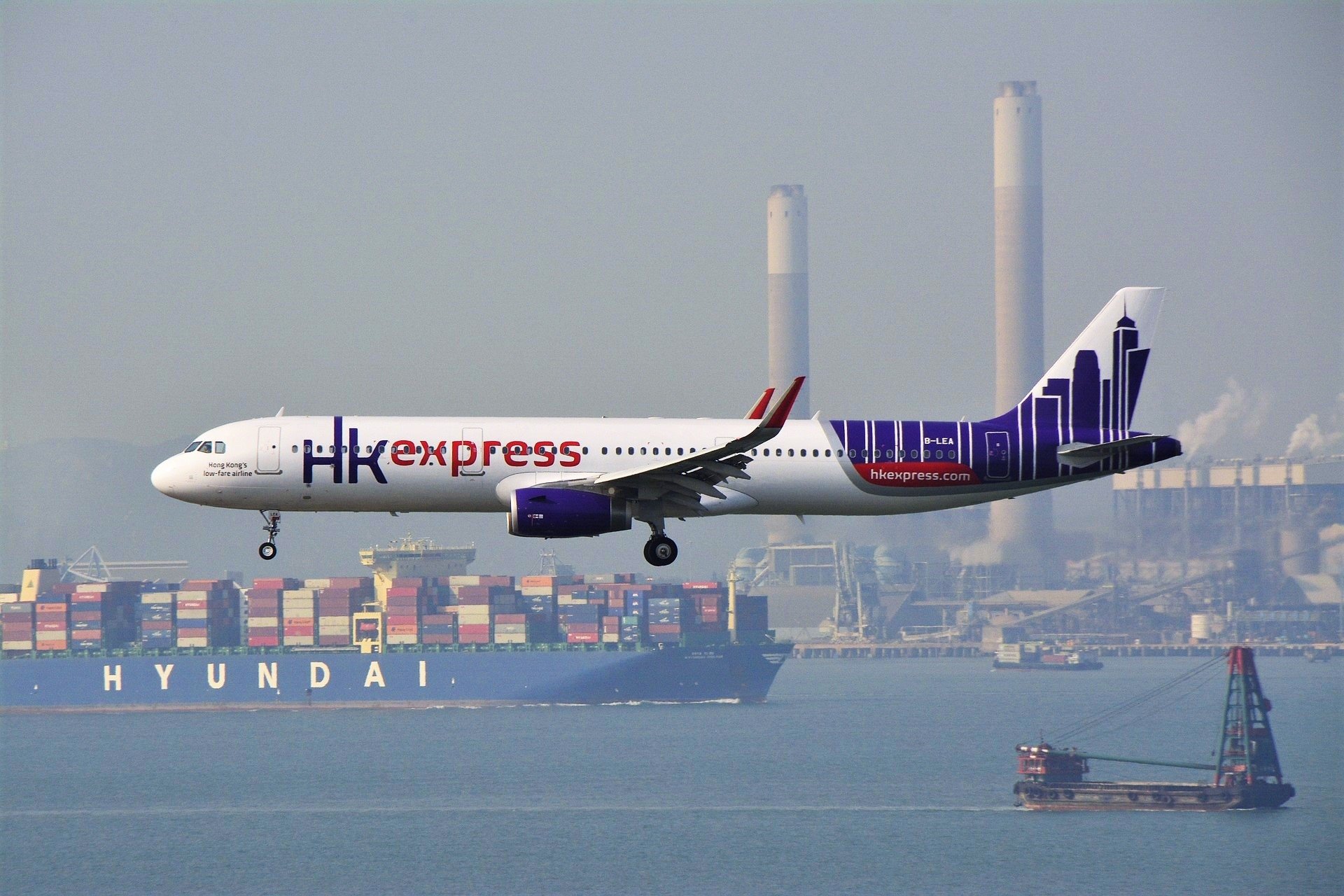 An Airbus A321 from HK Express approaches Hong Kong, with cargo ships and industrial structures visible in the background.