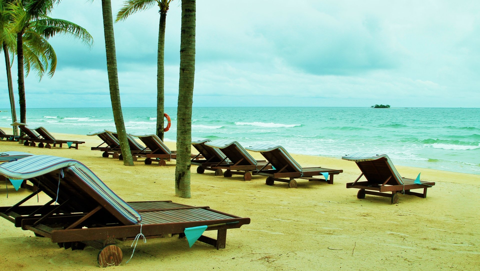 A row of beach chairs sits on a sandy shore, framed by palm trees, with a tranquil horizon of turquoise waters and a hint of clouds above, capturing the essence of a tropical holiday.