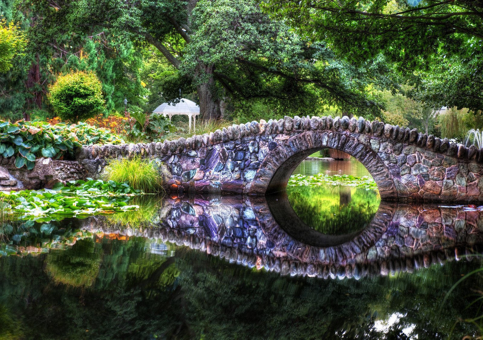 A picturesque stone bridge arches over a serene pond, surrounded by lush greenery and flowering plants, reflecting beautifully in the still water.