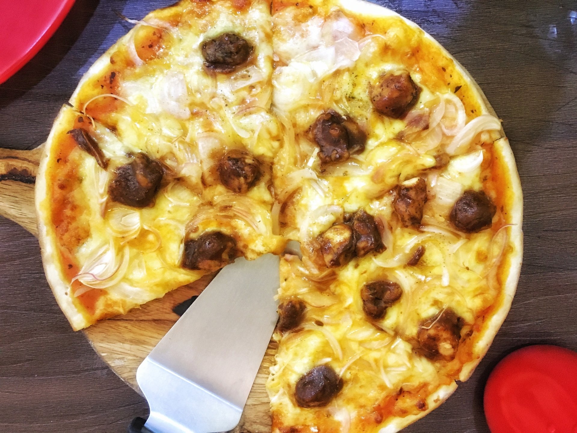 Food: a pizza on a wooden board with melted cheese, sliced onions and meatball-like toppings; one slice partially removed with a spatula.