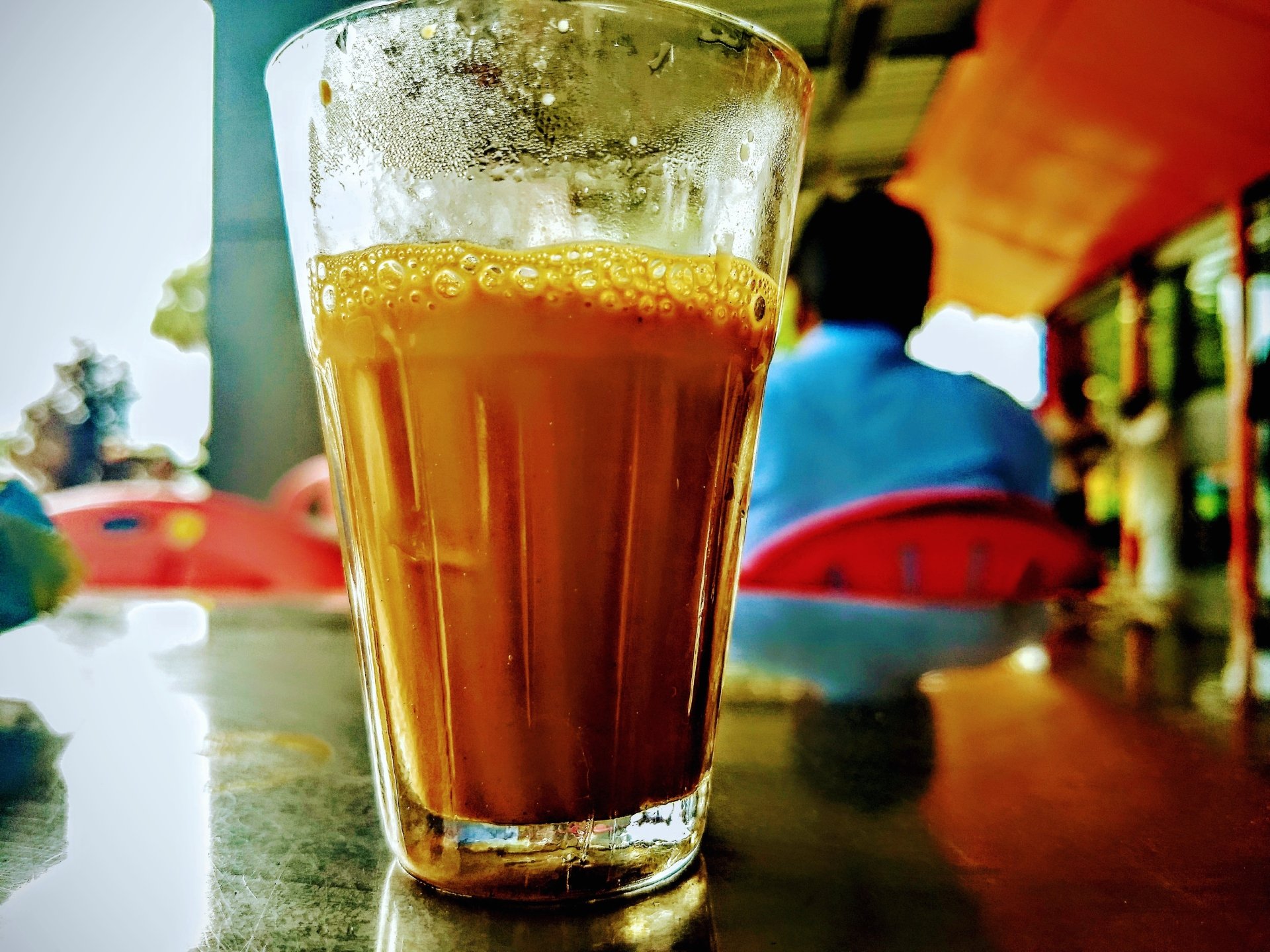 A glass of tea sits on a table, showcasing its rich brown color and frothy top, with a blurred figure in blue seated in the background. The scene captures a casual dining atmosphere.