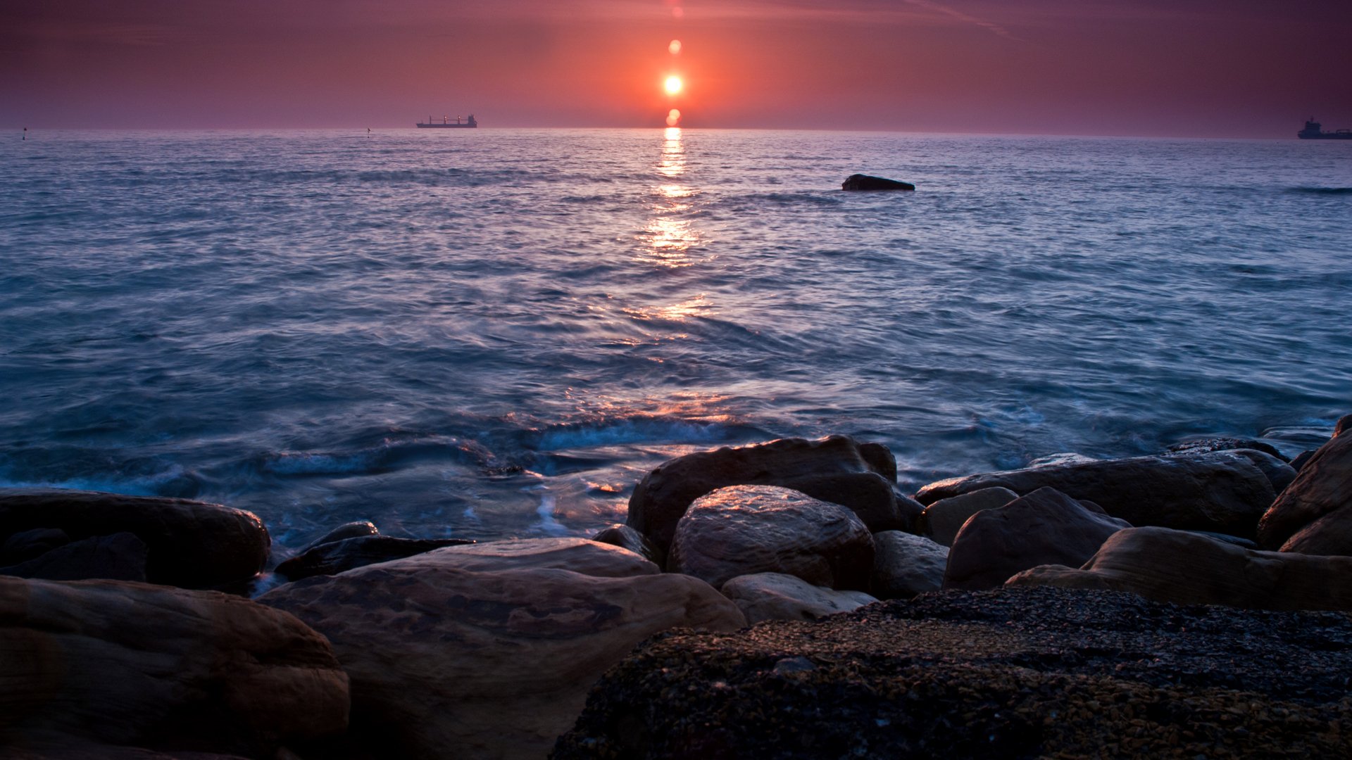 A serene sunset casts warm colors over the ocean, while gentle waves lap against the rocky shoreline, creating a tranquil nature scene.