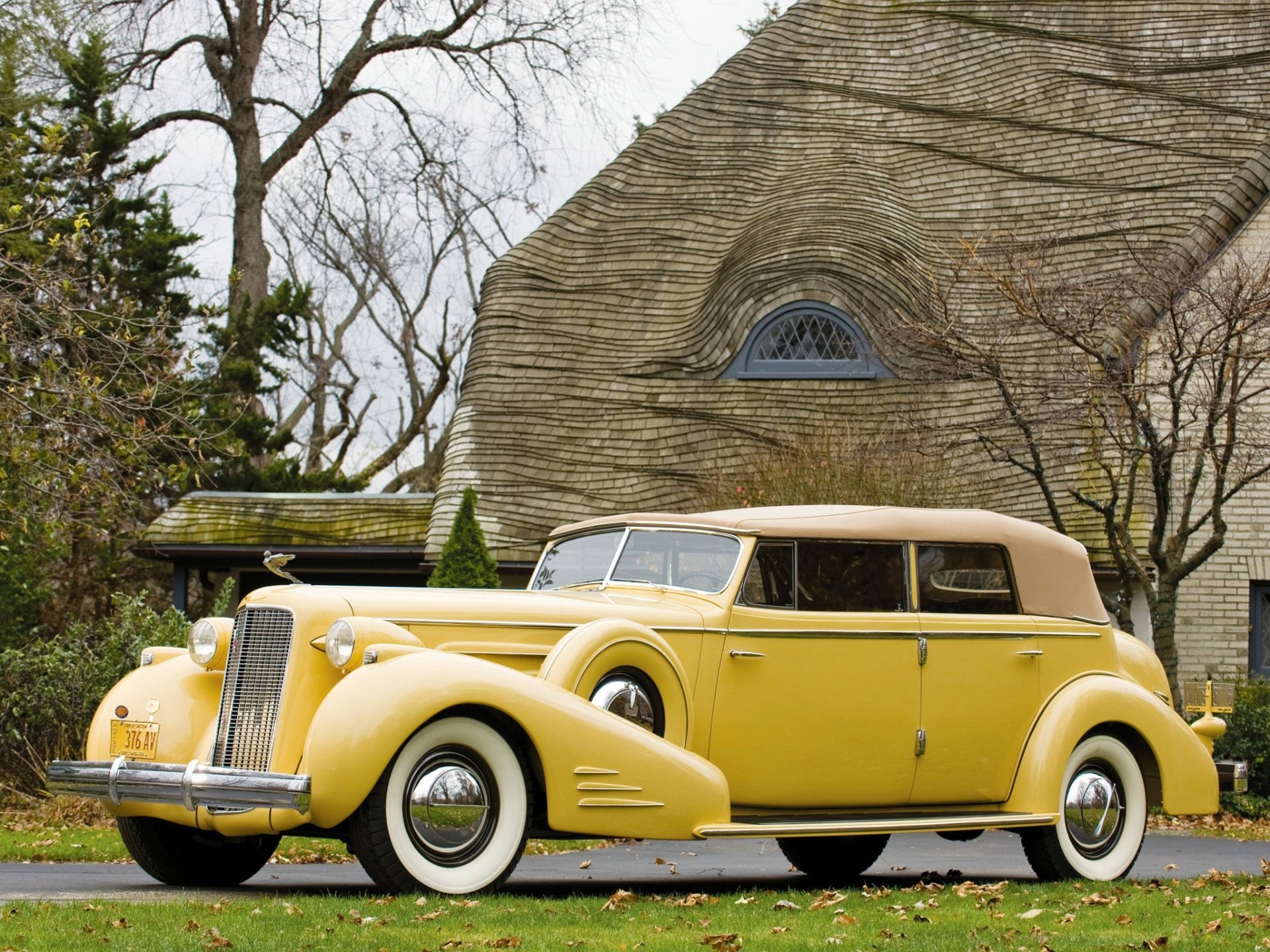 Yellow vintage Cadillac V-16 convertible with tan soft top parked on a lawn in front of a stone building and leafless trees.