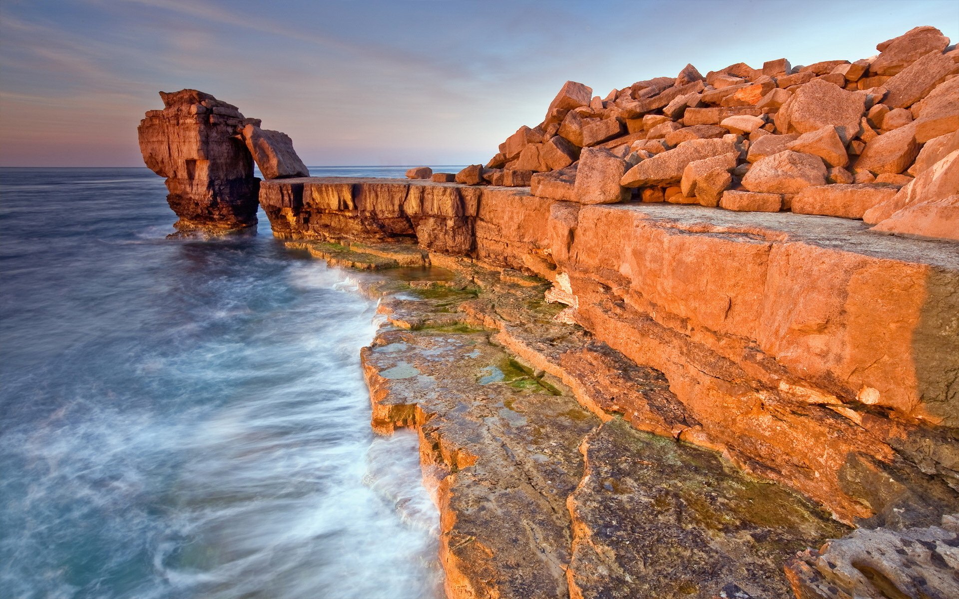 A rugged cliff juts out over the ocean, with golden light illuminating the rocks and waves crashing below, creating a stunning natural landscape.