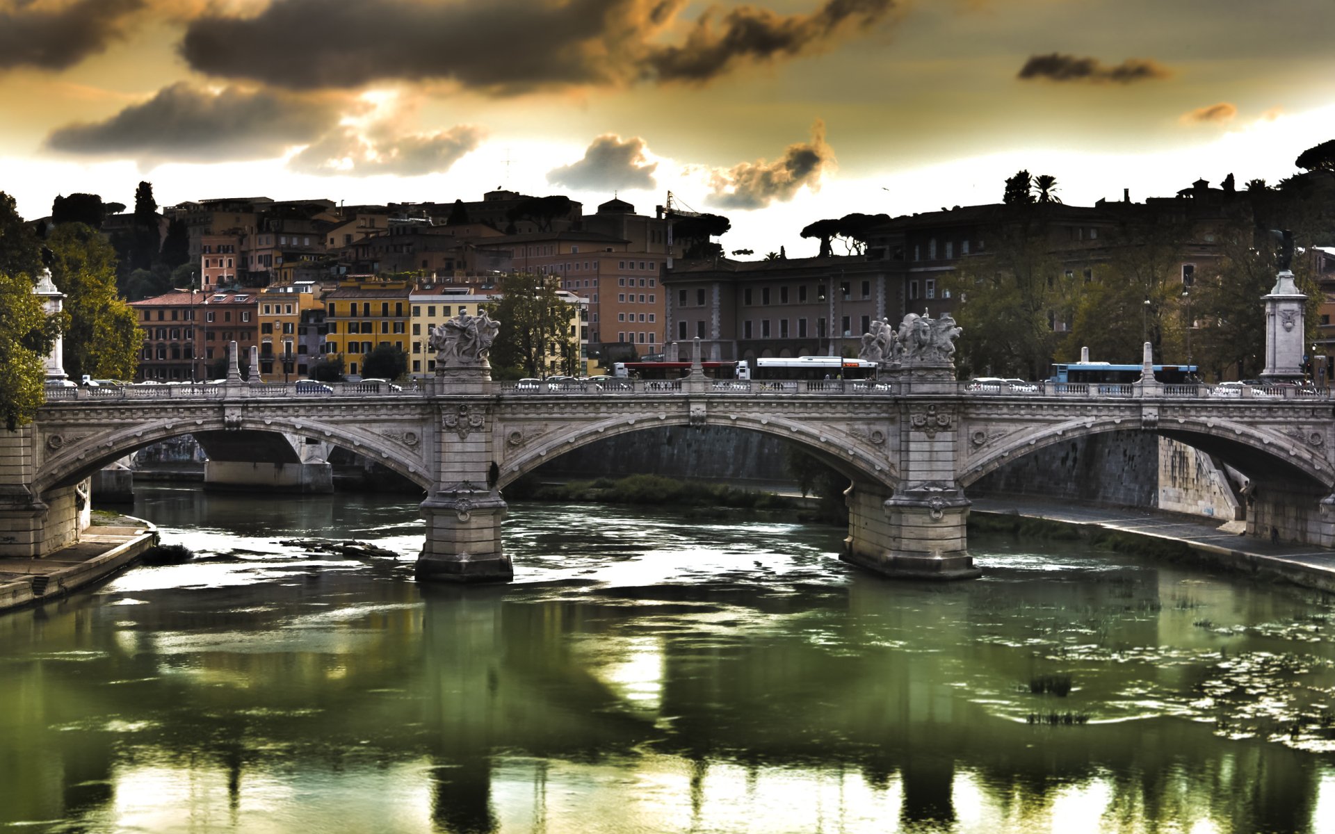 The Ponte Vittorio Emanuele II bridge spans a river with a cityscape and dramatic cloudy sky in the background.