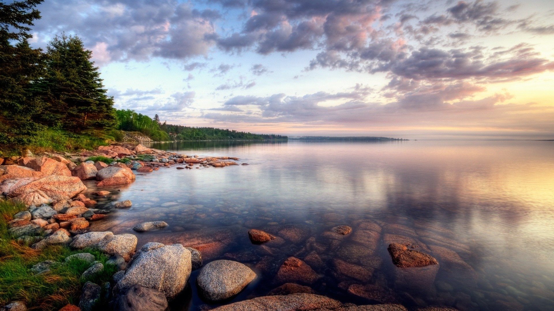 A tranquil coastline scene featuring smooth stones along the shore, with lush trees and a calm water surface reflecting the colorful sunset sky.