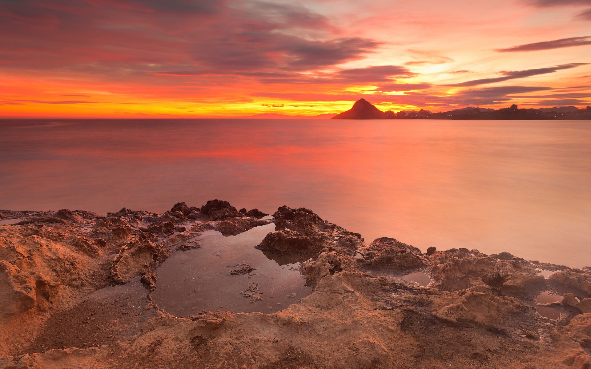 A vibrant nature scene capturing a sunset over calm waters with rocky formations in the foreground and a distant silhouetted mountain under a colorful sky.