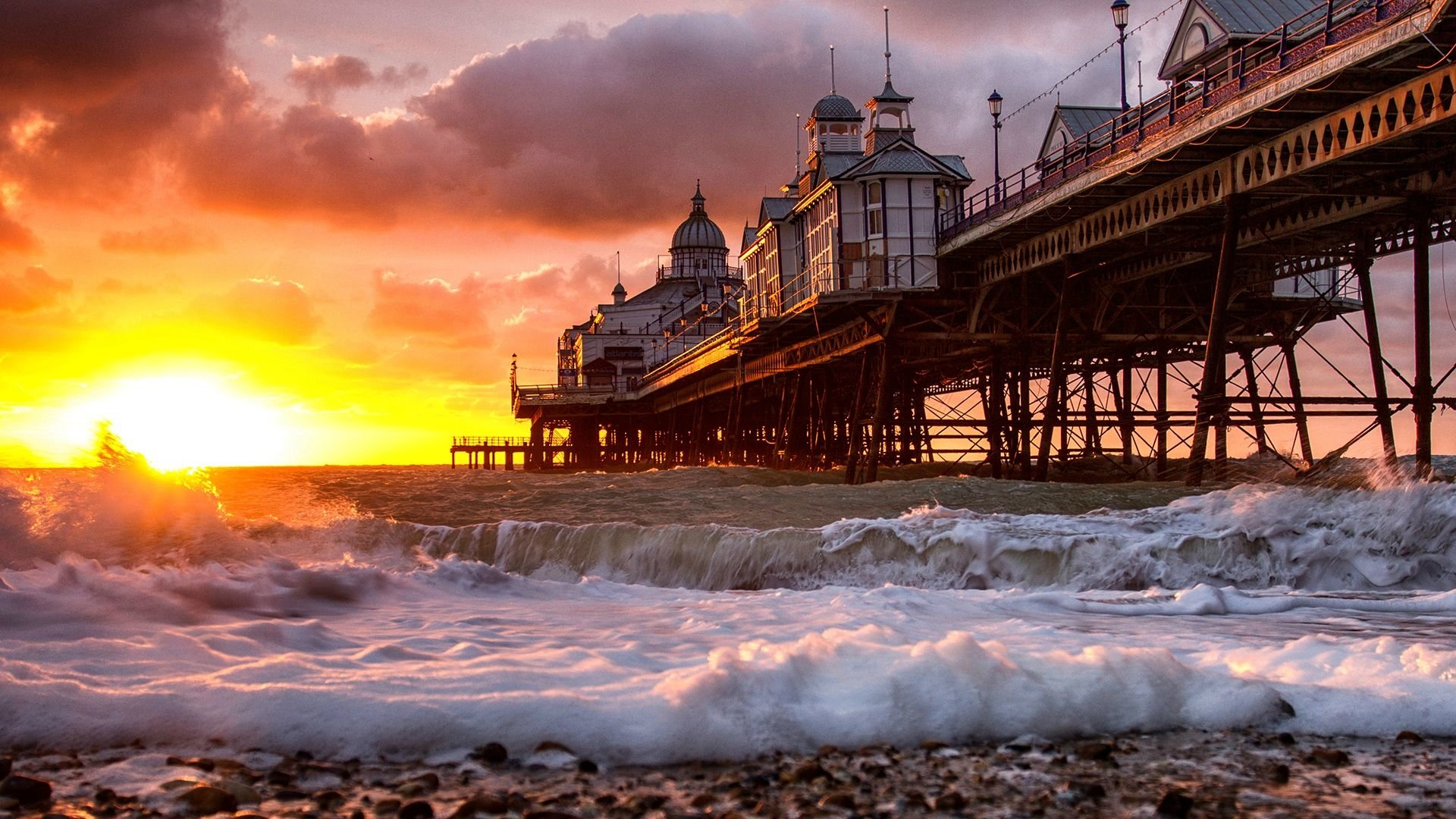 A vibrant sunset illuminates a manmade pier, creating a dramatic scene with waves crashing against its structure, accentuating the beauty of the coastline.