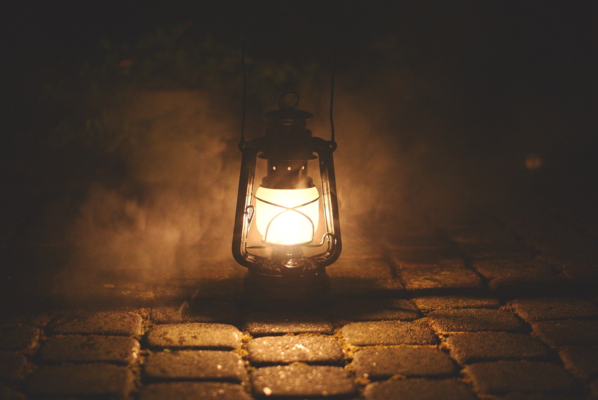 Warm glowing man-made lantern lamp casting light across damp cobblestones in a foggy night.