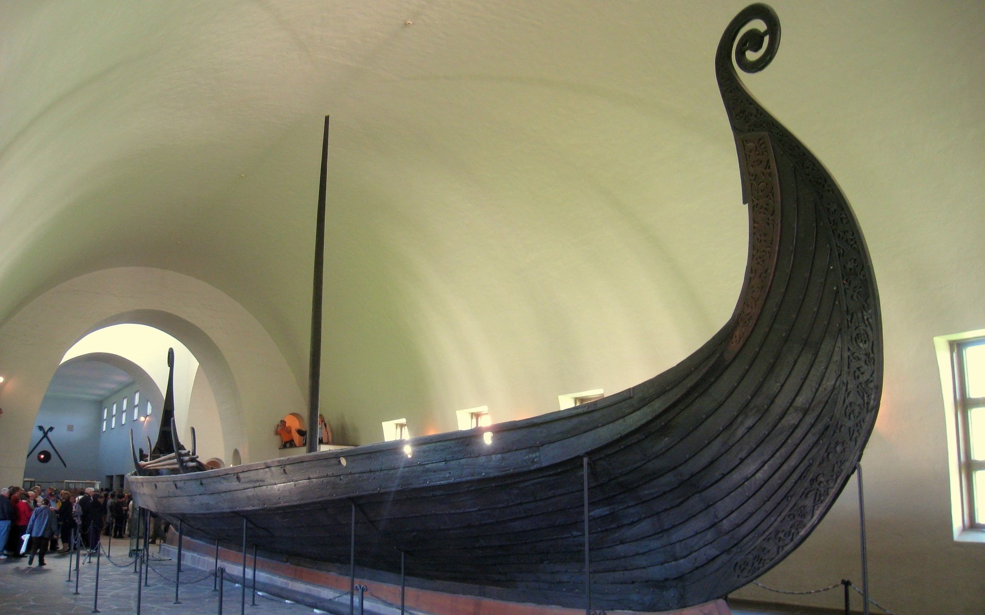 A large wooden drakkar, a traditional Viking ship, displayed inside a museum with visitors observing it from behind a barrier.