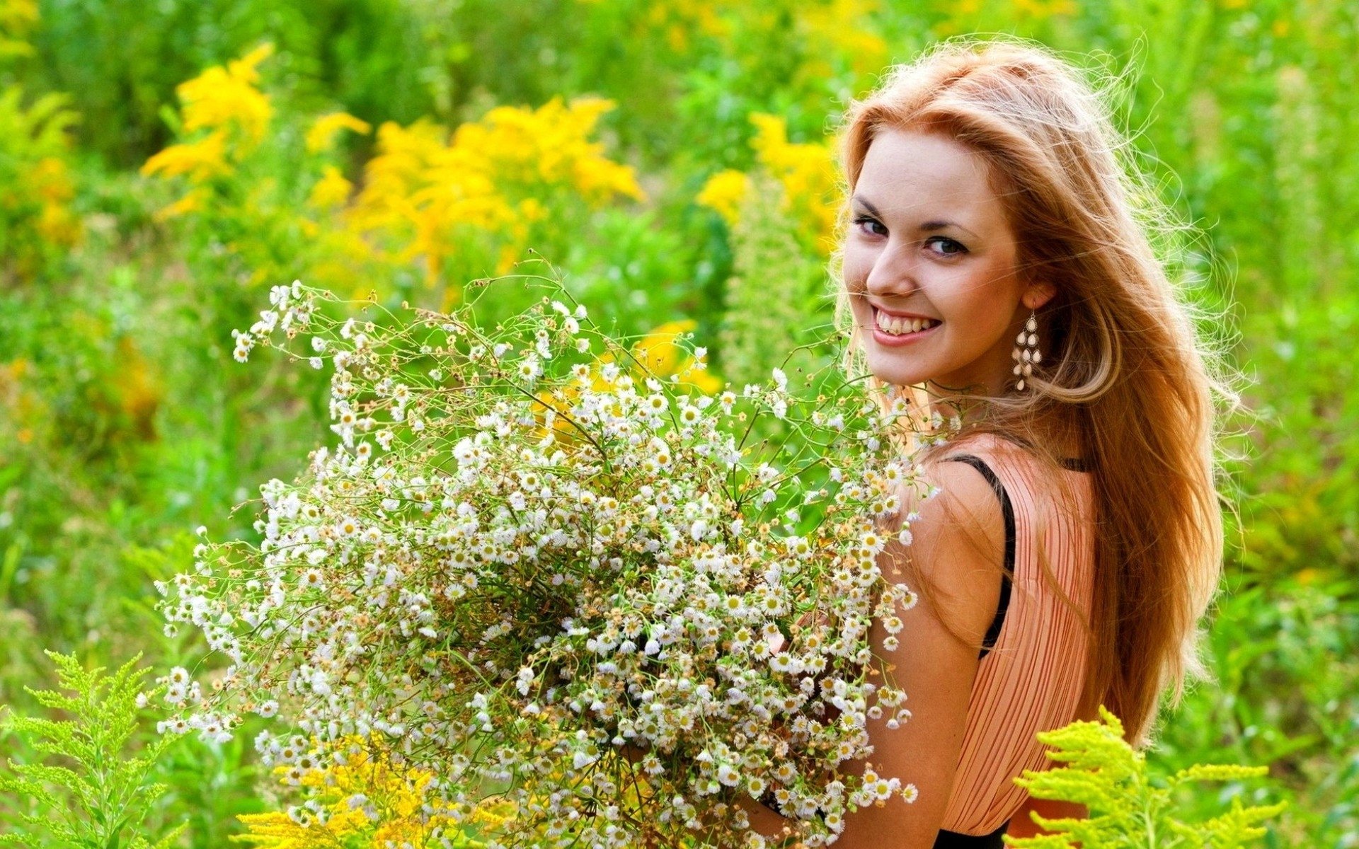 A woman smiles warmly while holding a large bouquet of wildflowers in a vibrant summer meadow, radiating a joyful and carefree mood.
