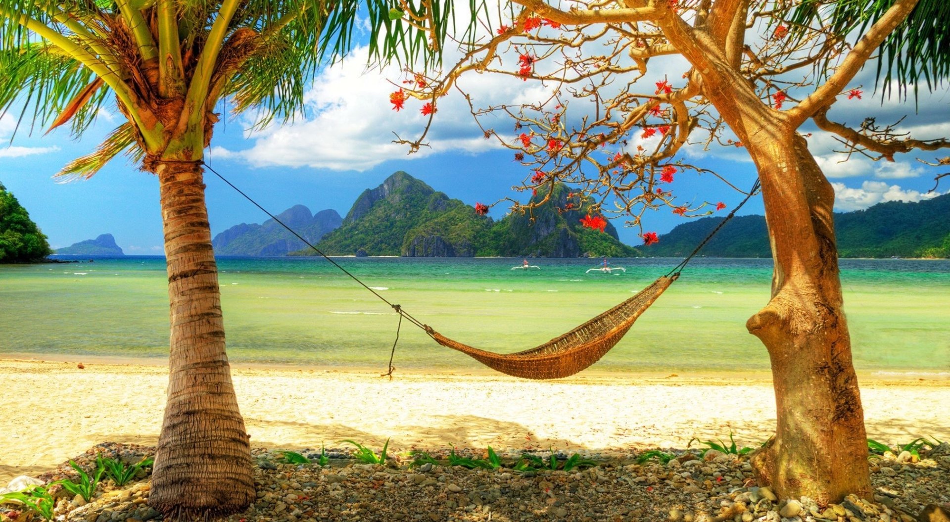 A tranquil tropical lagoon beach with a hammock strung between two palm trees, clear turquoise water, and lush green mountains in the background.