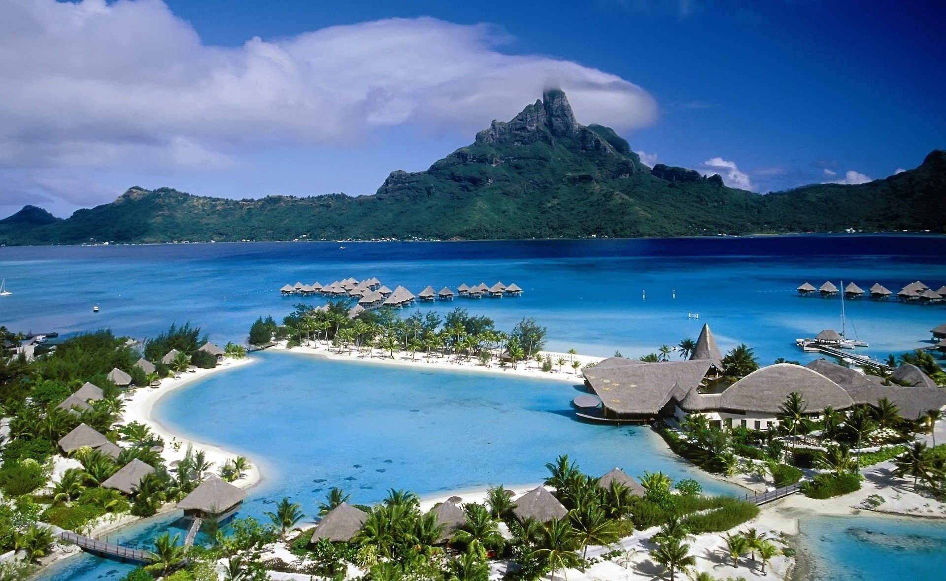 Tropical village nestled along a serene lagoon with overwater bungalows, set against a backdrop of lush mountains and deep blue sea under a partly cloudy sky.