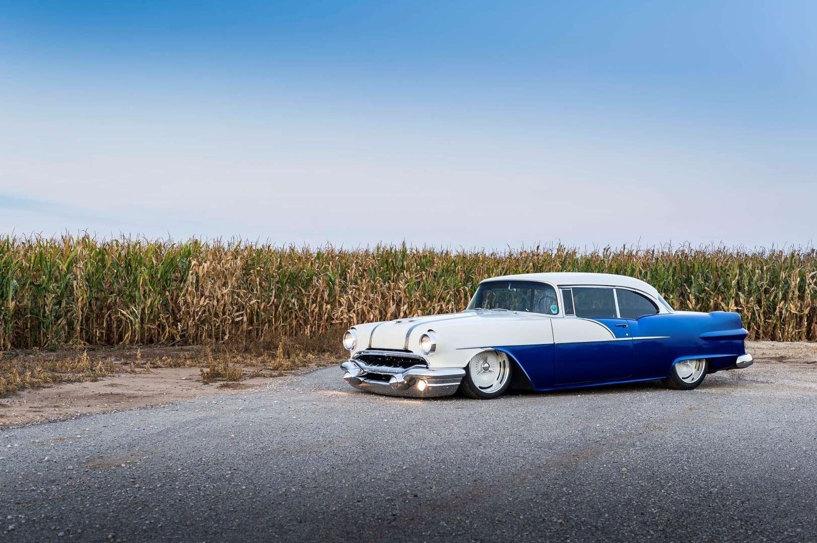A classic Pontiac Chieftain parked beside a cornfield, showcasing its striking blue and white paint, exuding vintage charm under a clear blue sky.