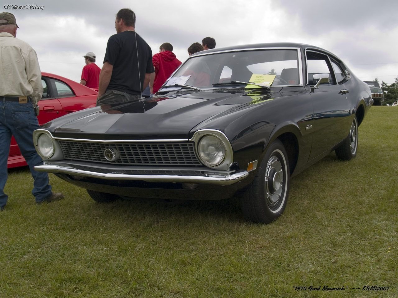 Classic dark green Ford Maverick vehicle parked on grass at a car show, people standing nearby and a paper notice tucked under the windshield wiper.