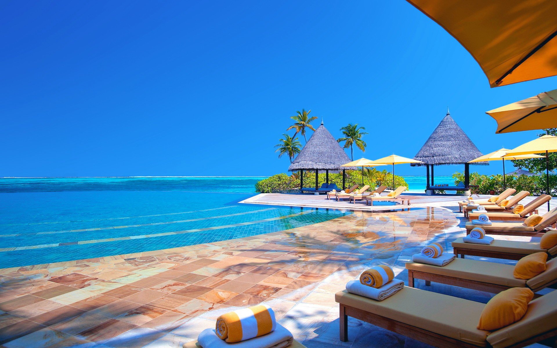 A tranquil resort scene with lounge chairs by a serene pool, surrounded by palm trees and thatched cabanas, set against a clear blue sky and sparkling water.