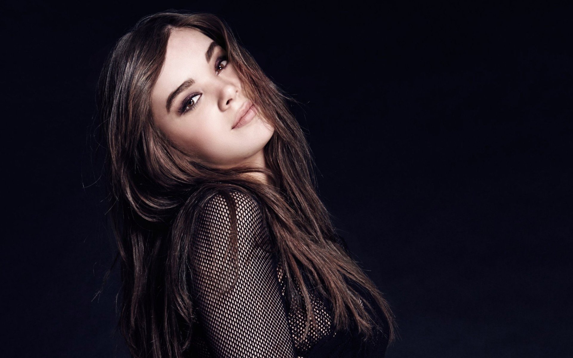 Hailee Steinfeld, a brunette American actress with striking brown eyes, poses confidently against a dark background, showcasing her charisma and style.