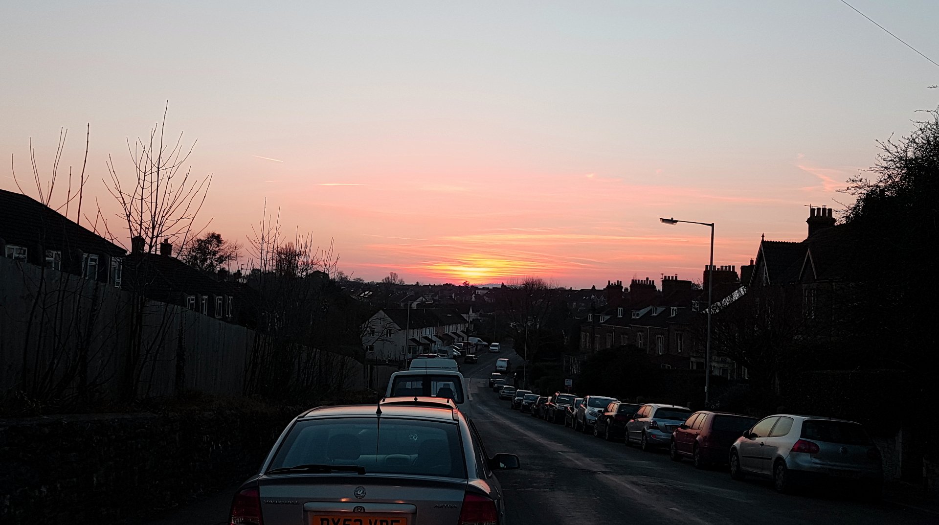 A serene twilight cityscape captures a stunning sunset, with soft hues of pink and orange painting the sky over a quiet street lined with parked cars.