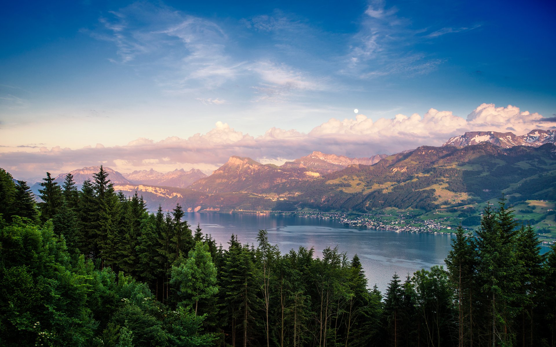 A serene view of a lush forest in the foreground, leading to majestic mountains and a tranquil lake, bathed in soft, natural light under a clear blue sky.