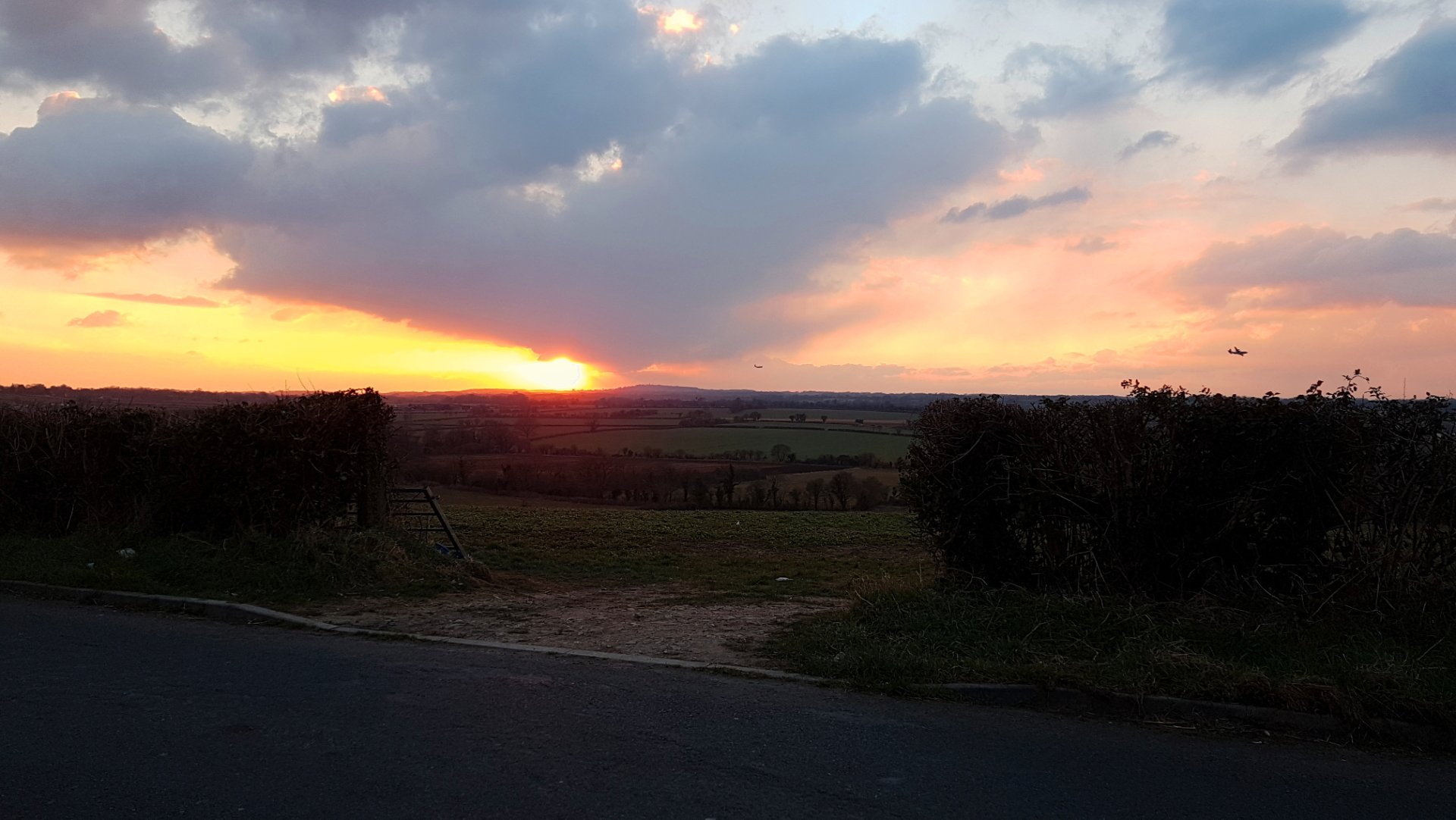 Roadside hedgerow opening to rolling fields under a dramatic cloud-filled sky at sunset, warm orange sun near the horizon — landscape photography.