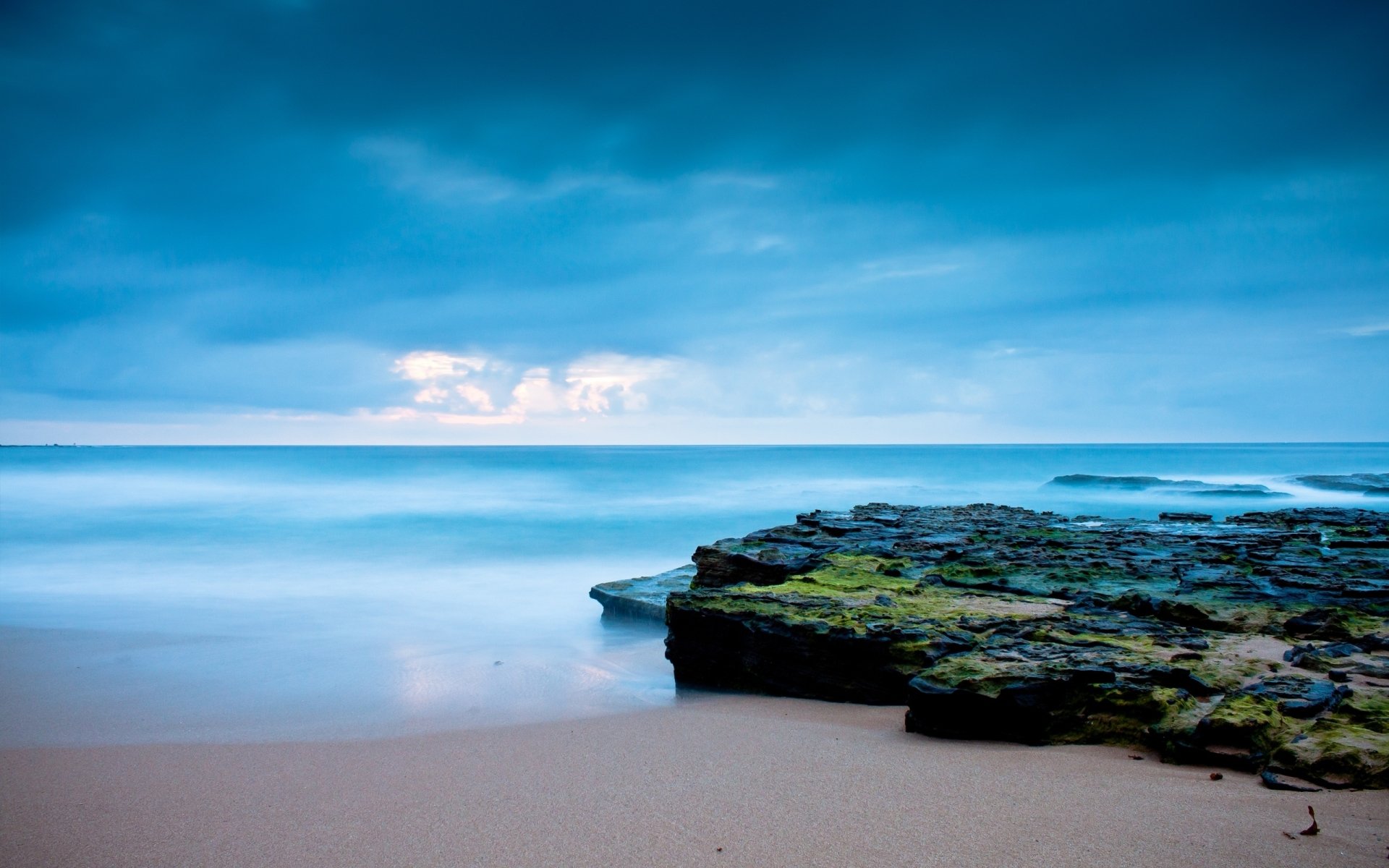 Calm ocean waves gently meet a rocky shoreline under a vast blue sky with a distant horizon stretching across the serene natural scene.