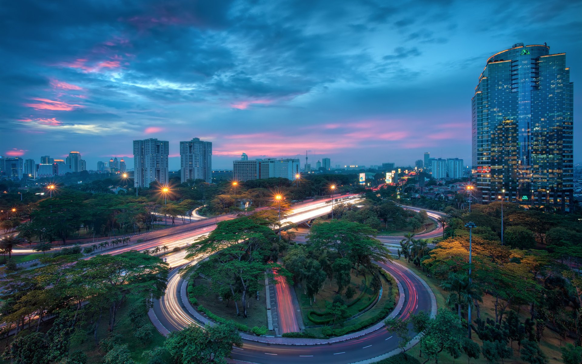 Sunset over Jakarta cityscape in Indonesia, featuring illuminated roads, greenery, and tall buildings beneath a colorful sky.