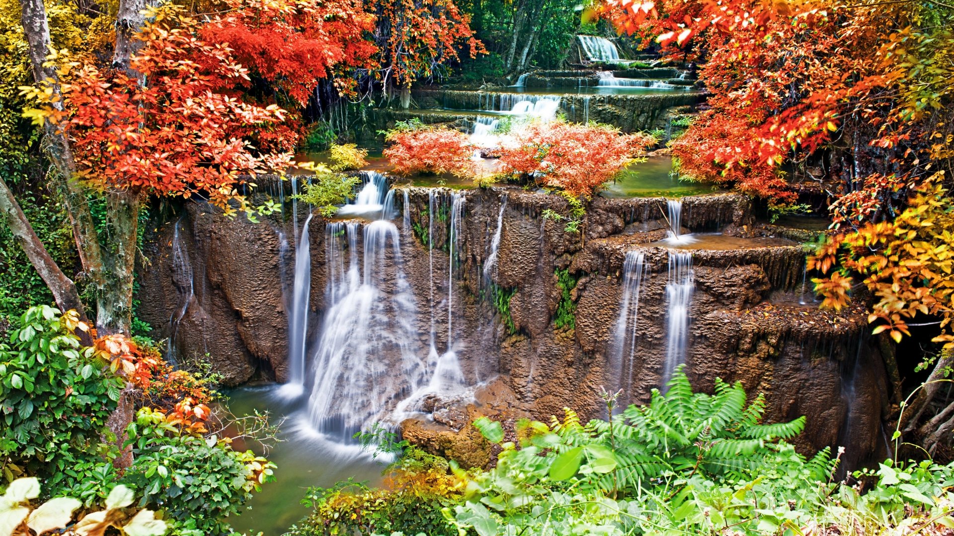 A vibrant waterfall cascades down rocky ledges, surrounded by lush greenery and trees in autumn hues of orange and red, showcasing the beauty of nature.