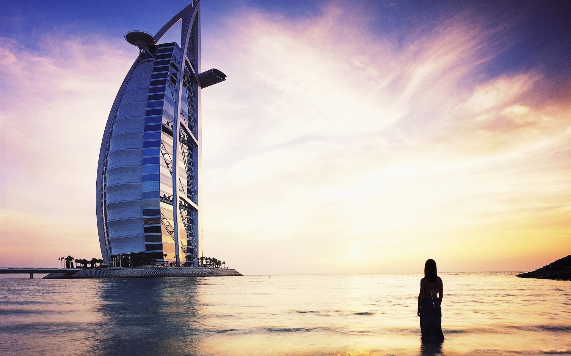 Silhouette of a person standing in shallow water at sunset with the man-made Burj Al Arab hotel towering to the left beneath a vivid, colorful sky.