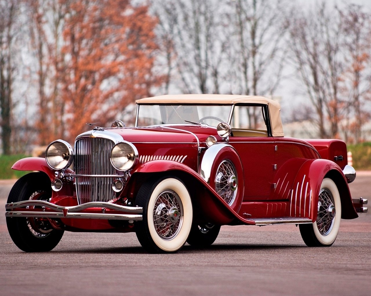 Maroon Duesenberg Model J convertible with tan soft top, chrome grille, whitewall tires and wire-spoke wheels parked on pavement with bare trees in the background.