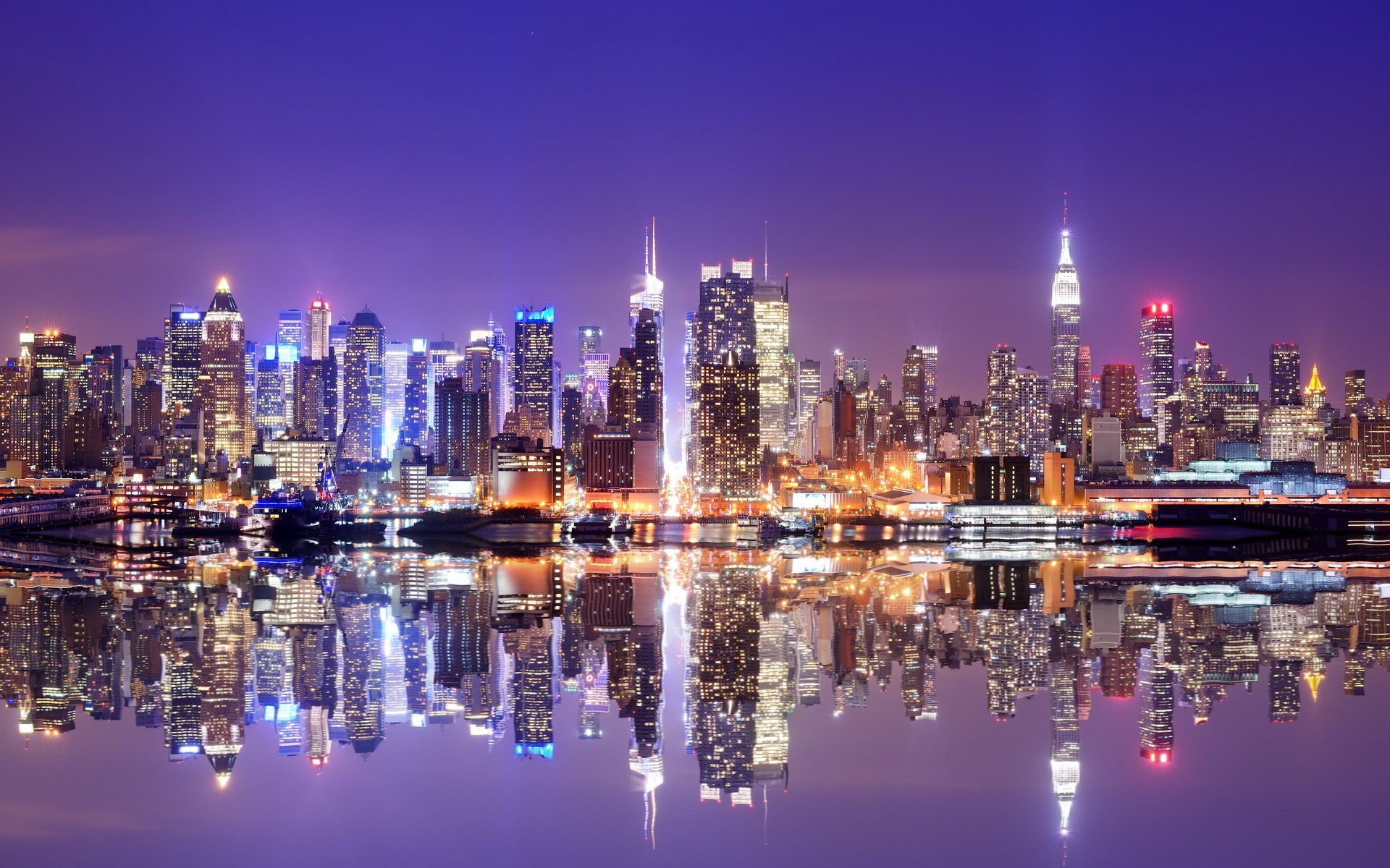A stunning night view of Manhattan, showcasing its iconic skyline reflected in calm waters, illuminated by vibrant city lights against a deep blue sky.