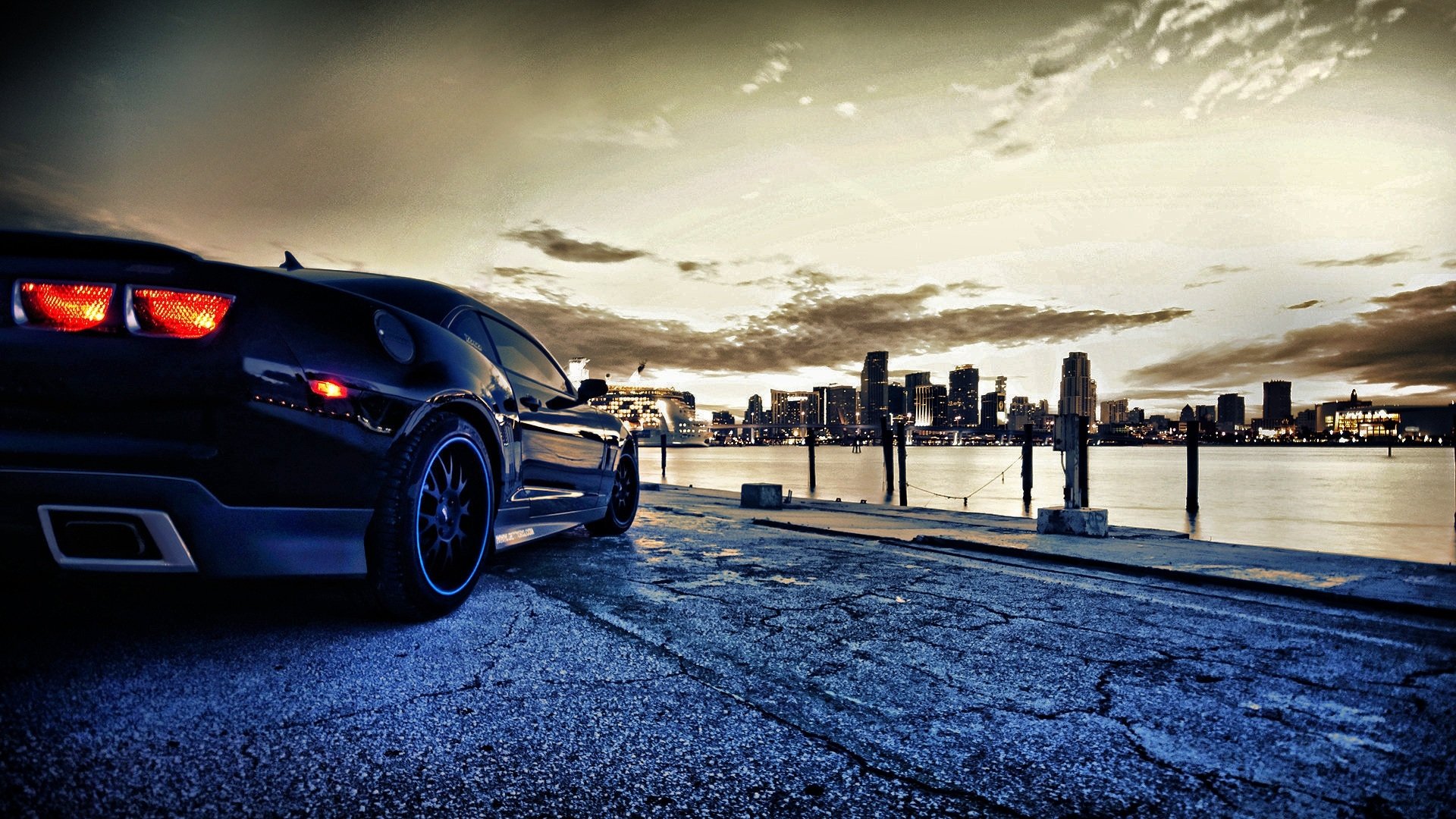 Black Chevrolet Camaro SS parked on a waterfront, Miami skyline and dramatic cloud-filled dusk sky reflected on the water.