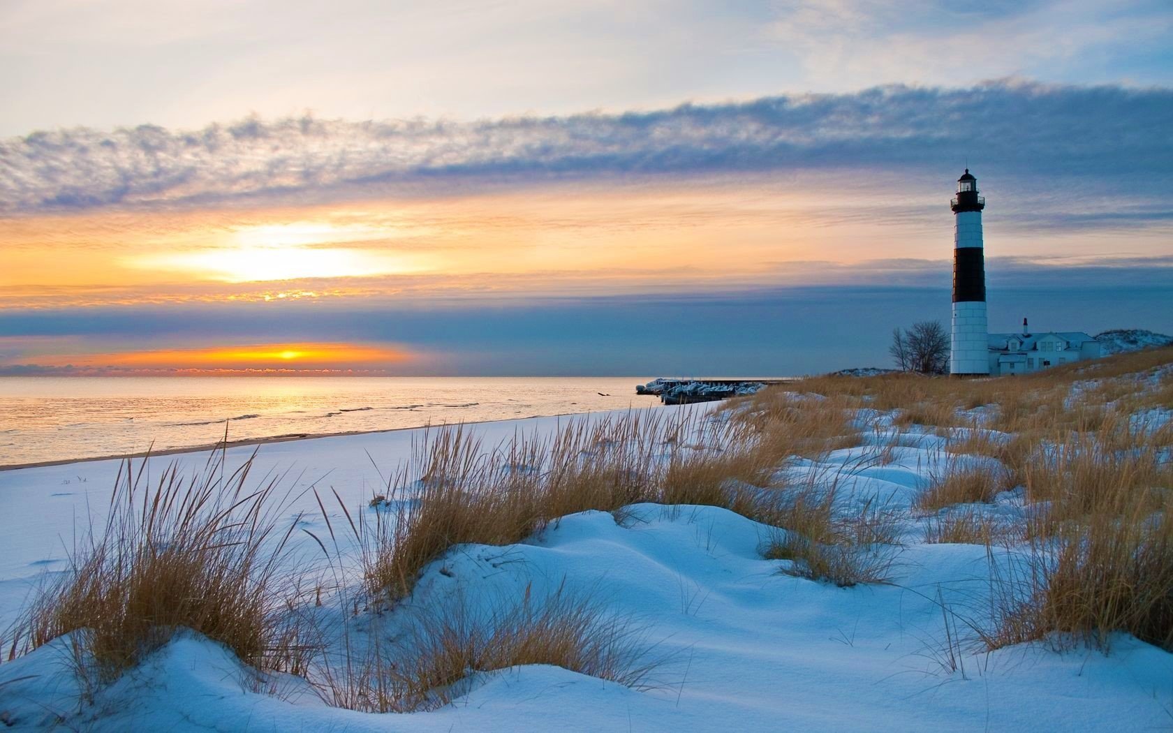 Snowy dune grasses and sand beside a calm sea at sunset on the horizon, with a black-and-white man-made lighthouse standing on the shore.
